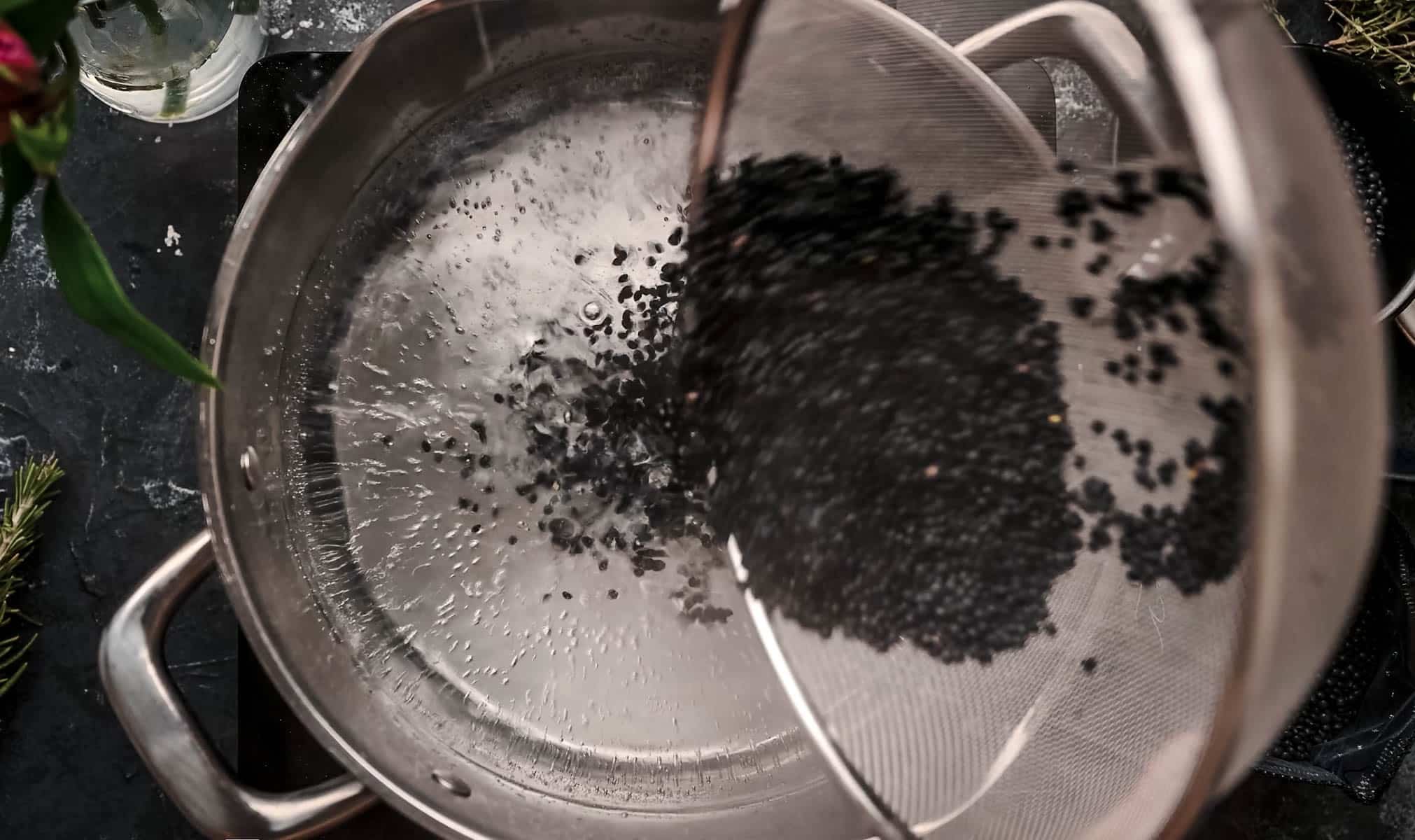 A sieve is used to pour black lentils into a stainless steel pot filled with water.