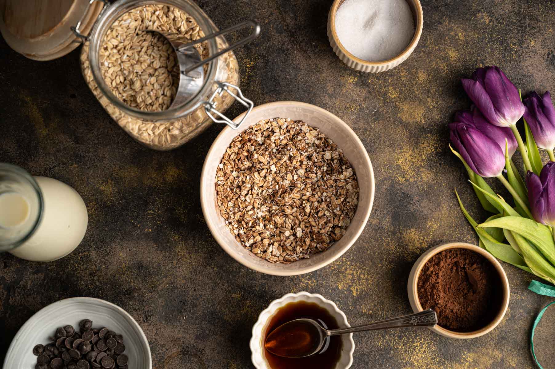 Top-down view of a kitchen countertop with oats in a bowl and jar, milk, chocolate chips, cocoa powder, maple syrup, and purple tulips arranged.