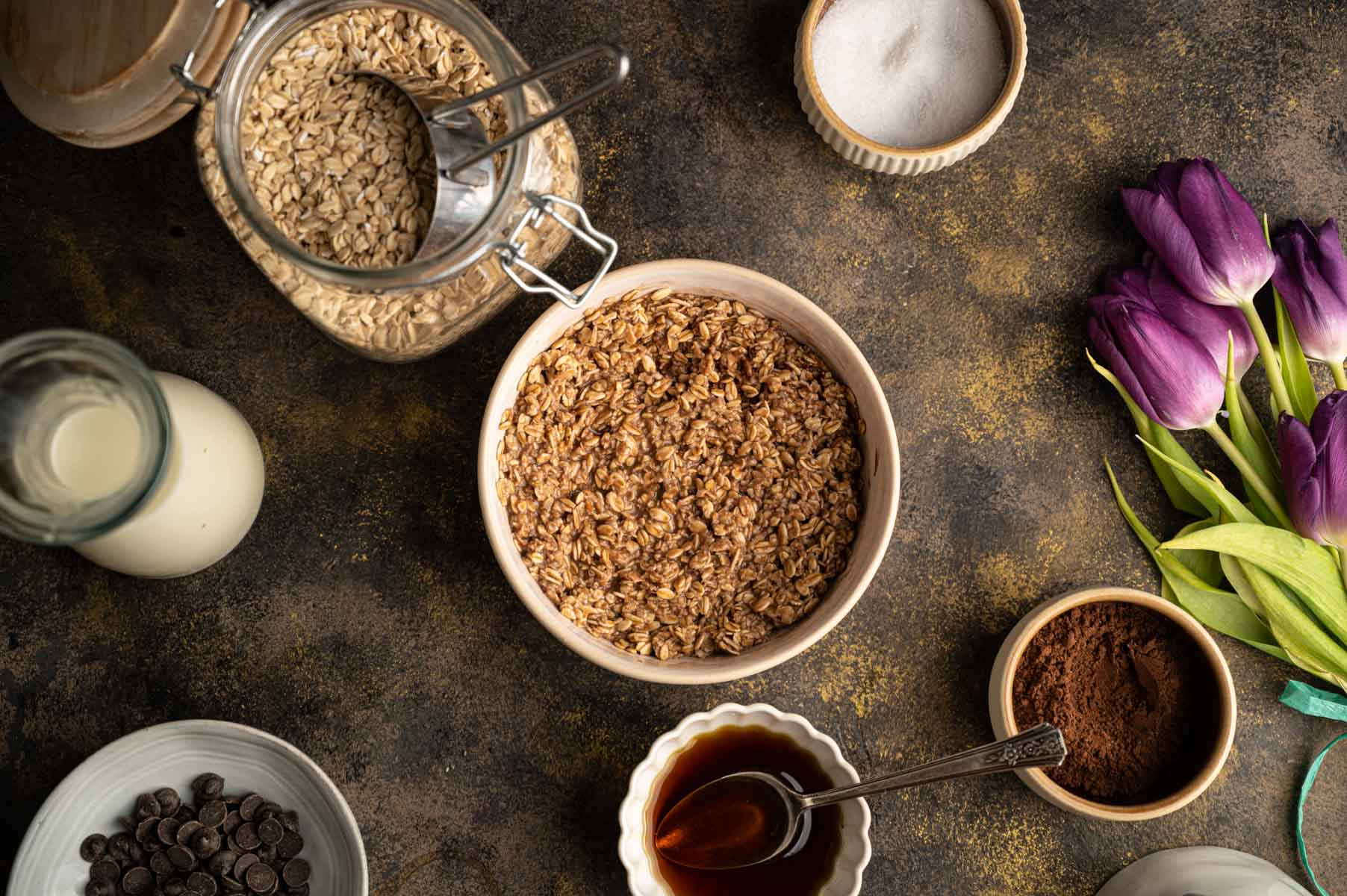 Top-down view of a kitchen counter with a bowl of oats, a jar of oats, plant milk, chocolate chips, and cocoa powder.