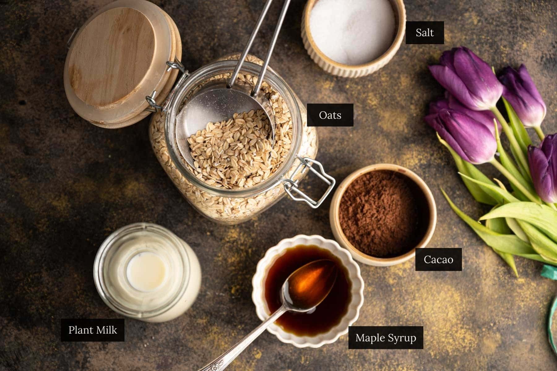 Overhead view of oats in a jar with a spoon, surrounded by bowls of salt, cacao, maple syrup, and a jar of plant milk. Purple tulips are placed to the right.