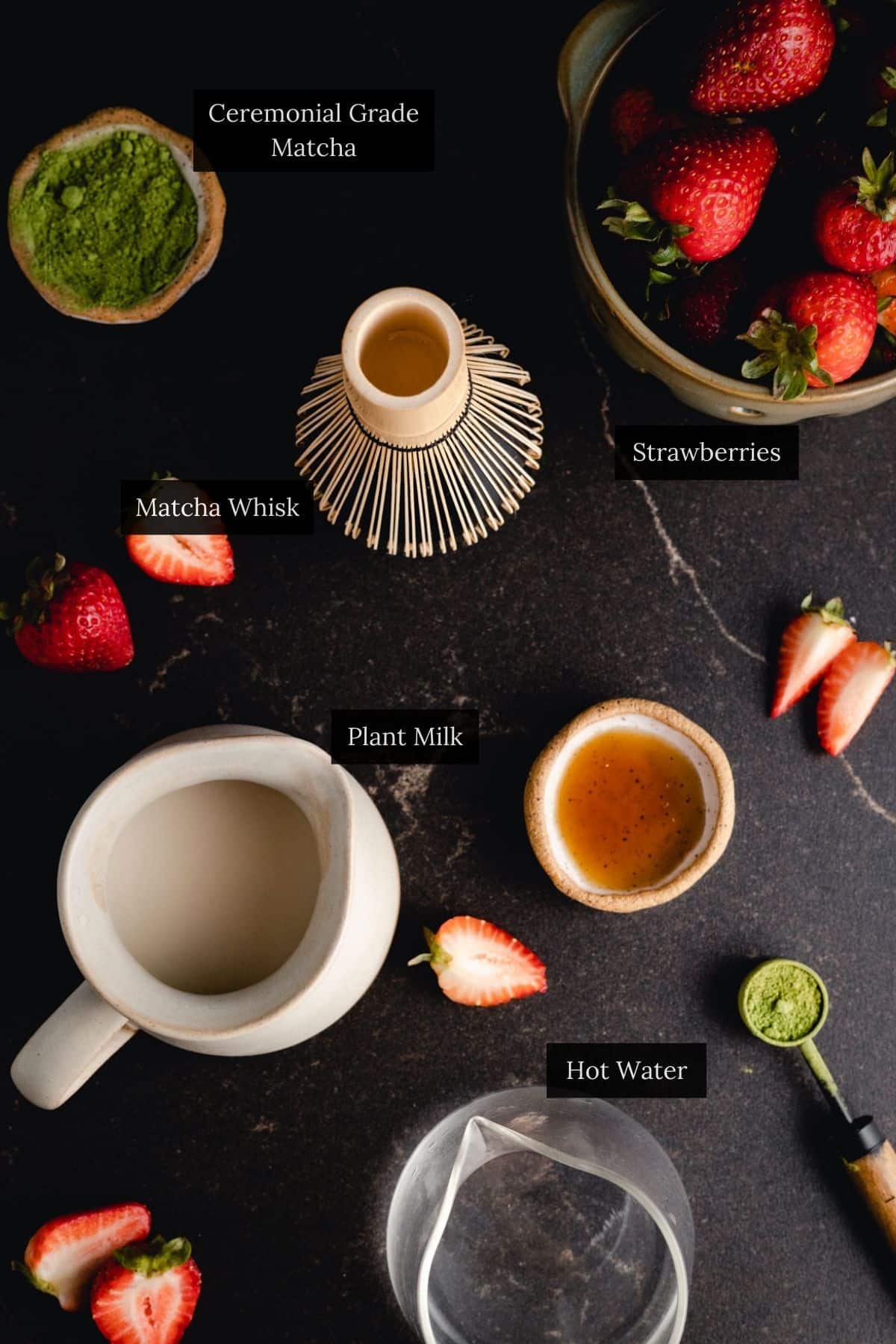 Top view of a dark surface showcasing a strawberry matcha latte setup, with fresh strawberries, matcha powder, a whisk, plant milk, hot water, and a small cup of vibrant green tea powder.