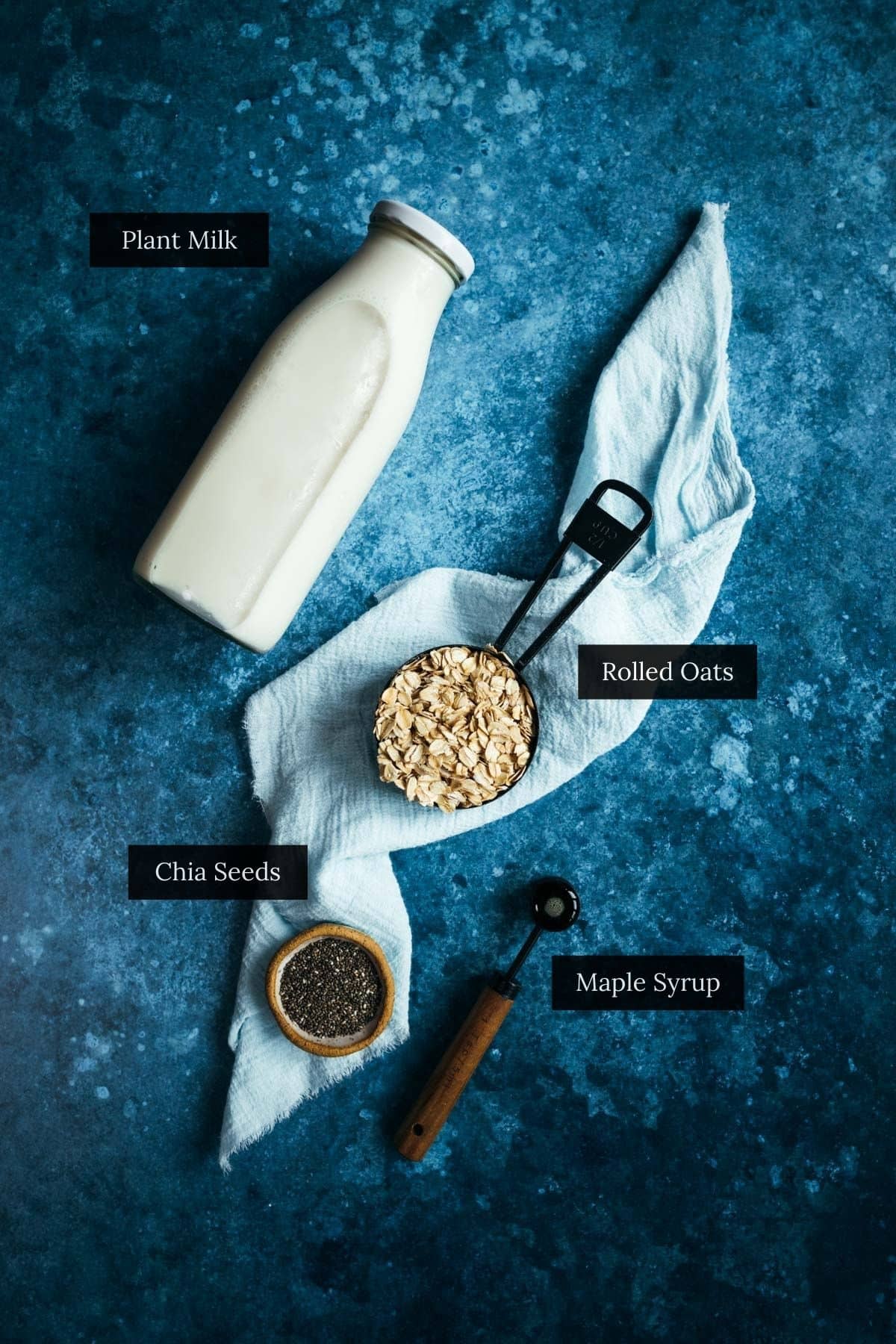A bottle of plant milk, a measuring cup with rolled oats, a small bowl of chia seeds, and a spoon with maple syrup arranged on a blue textured surface with a cloth napkin.