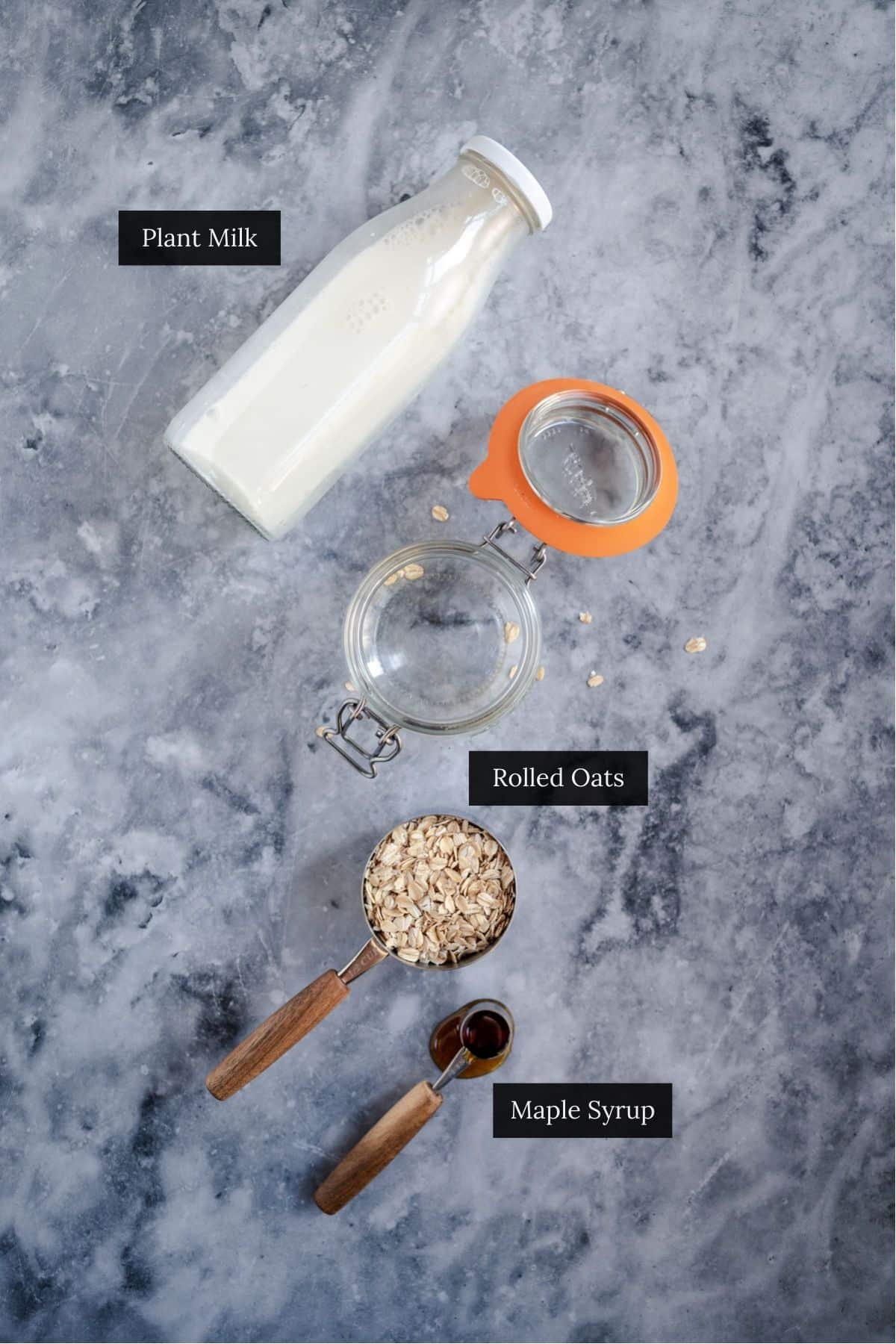 A bottle of plant milk, a jar of rolled oats, a measuring cup of rolled oats, and a spoon of maple syrup arranged on a marble surface.