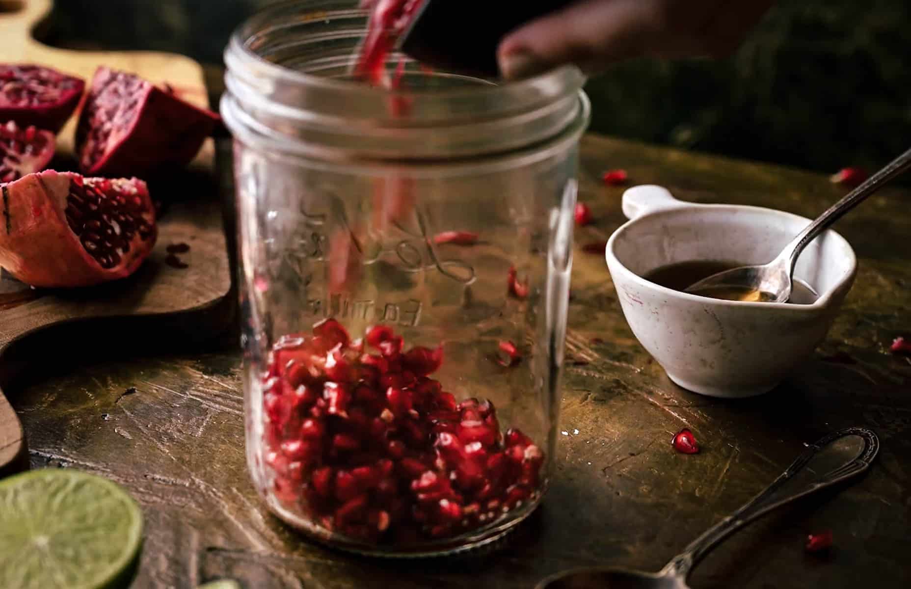 A person pours pomegranate seeds into a glass jar on a wooden surface. Nearby are a chopped pomegranate, lime, spoon, and small white cup.
