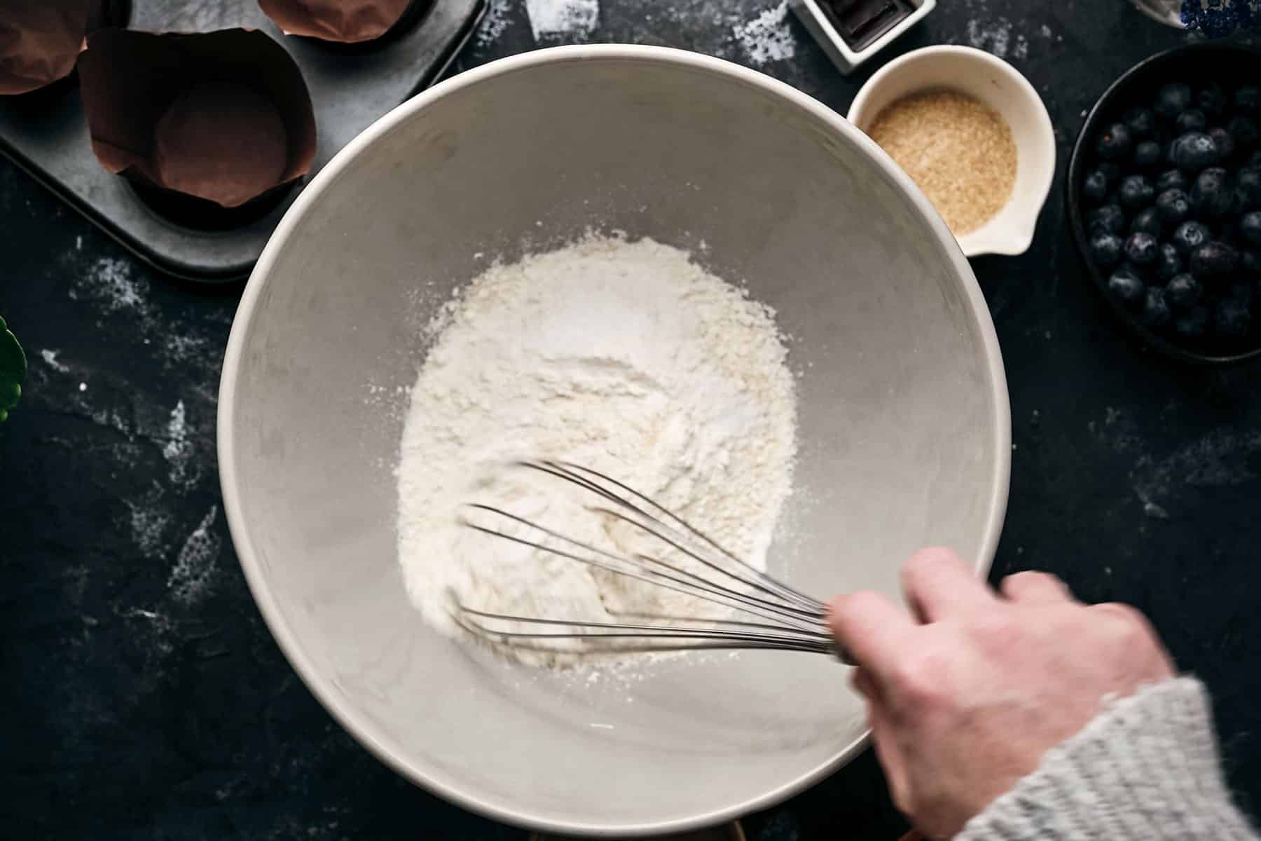 A person's hand is whisking flour in a large white bowl. Nearby, muffin liners, a small bowl of brown sugar, and blueberries are visible.