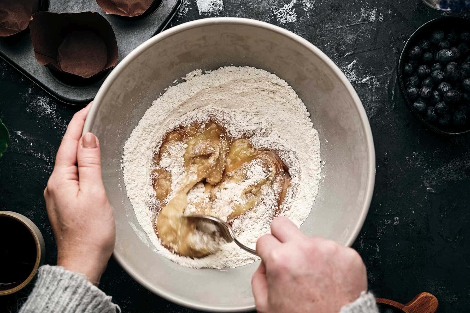 Hands mixing mashed bananas into flour in a large bowl.