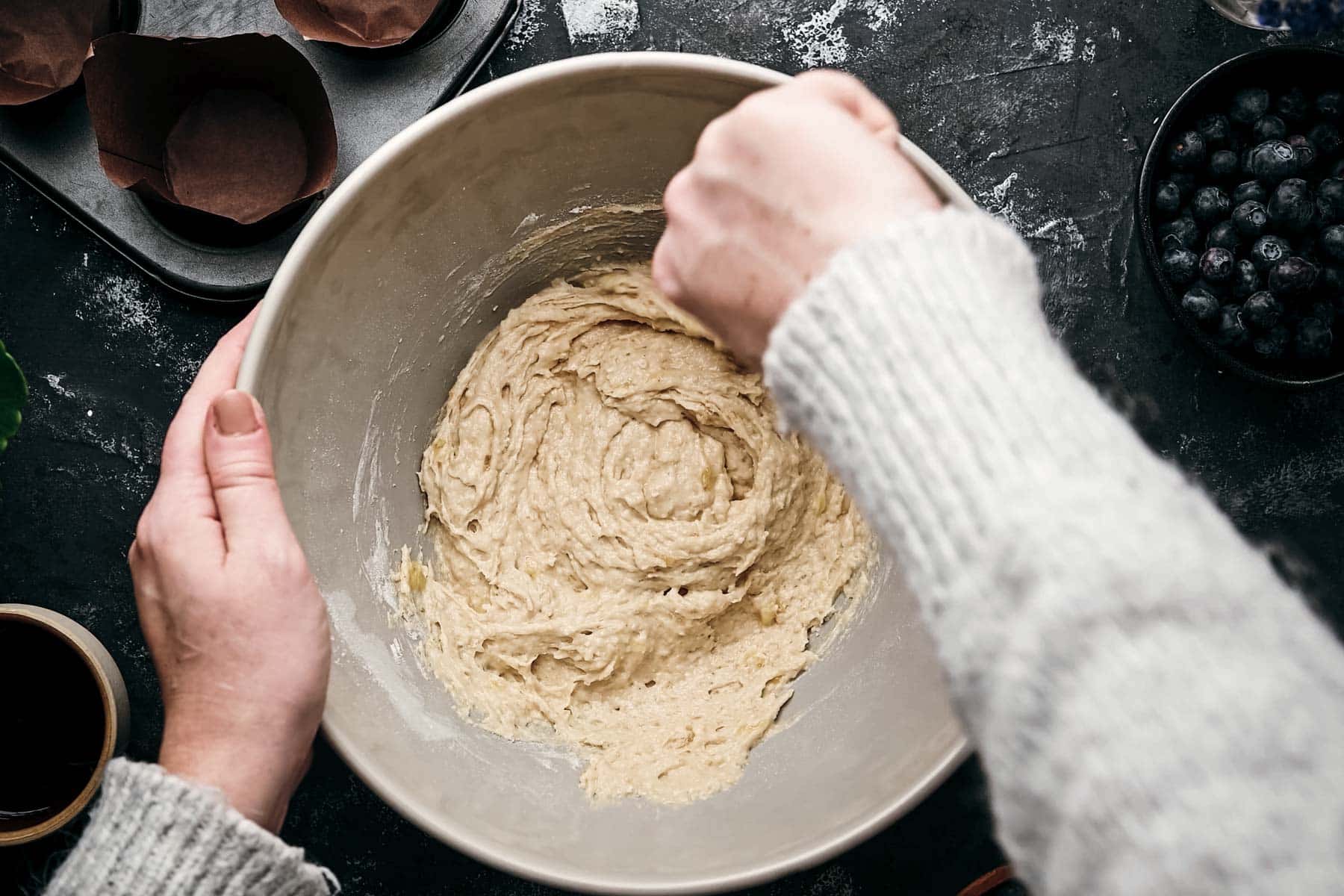 Hands stirring muffin batter in a mixing bowl, with blueberries and a muffin tray in the background.