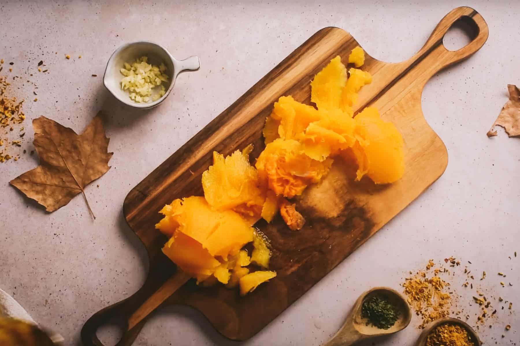 Chopped squash on a wooden cutting board, accompanied by a small bowl of minced garlic, dried leaves, and wooden spoons containing herbs and spices.