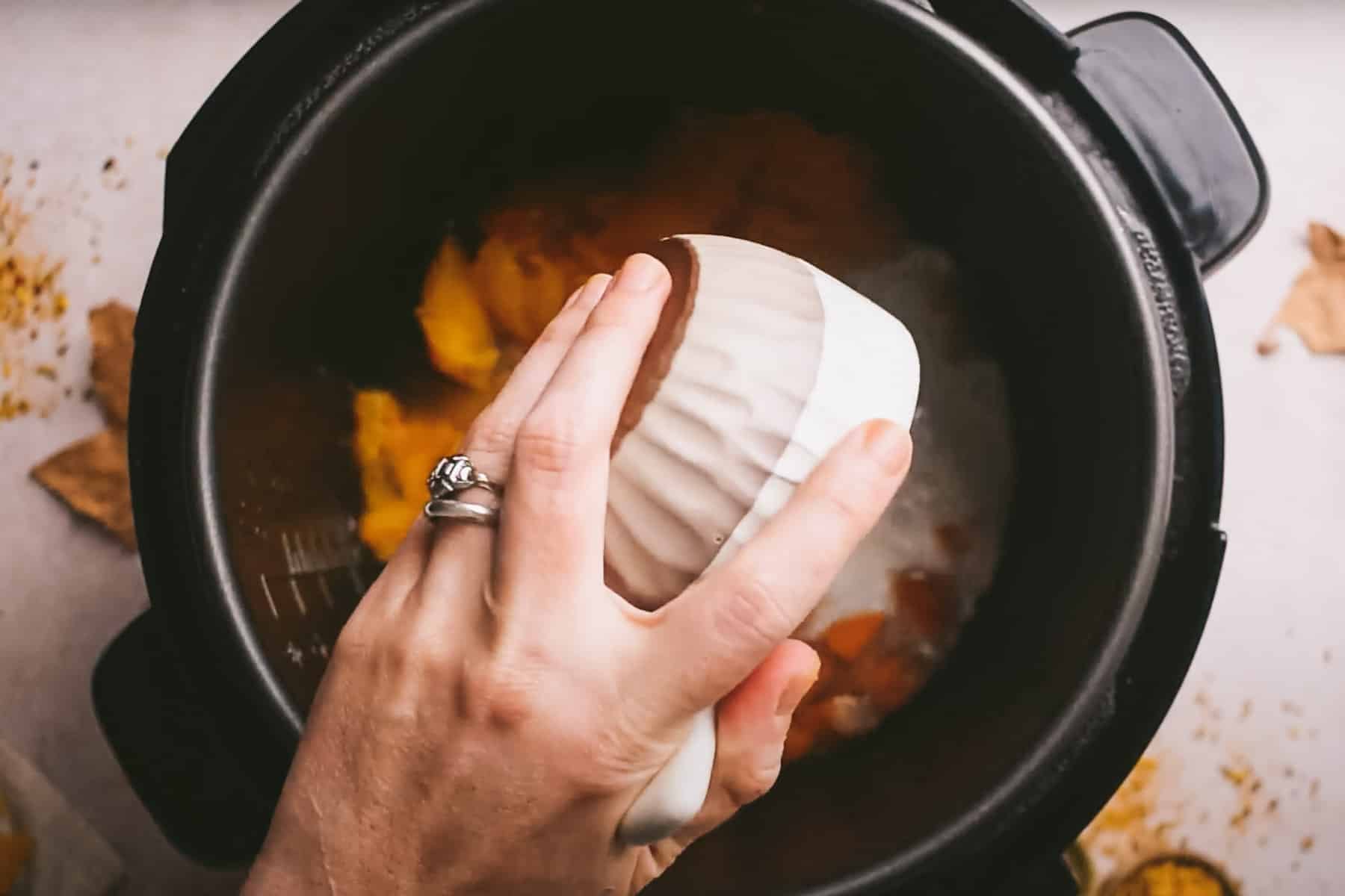 A hand with a ring is holding a white ceramic bowl above a black pot filled with food.