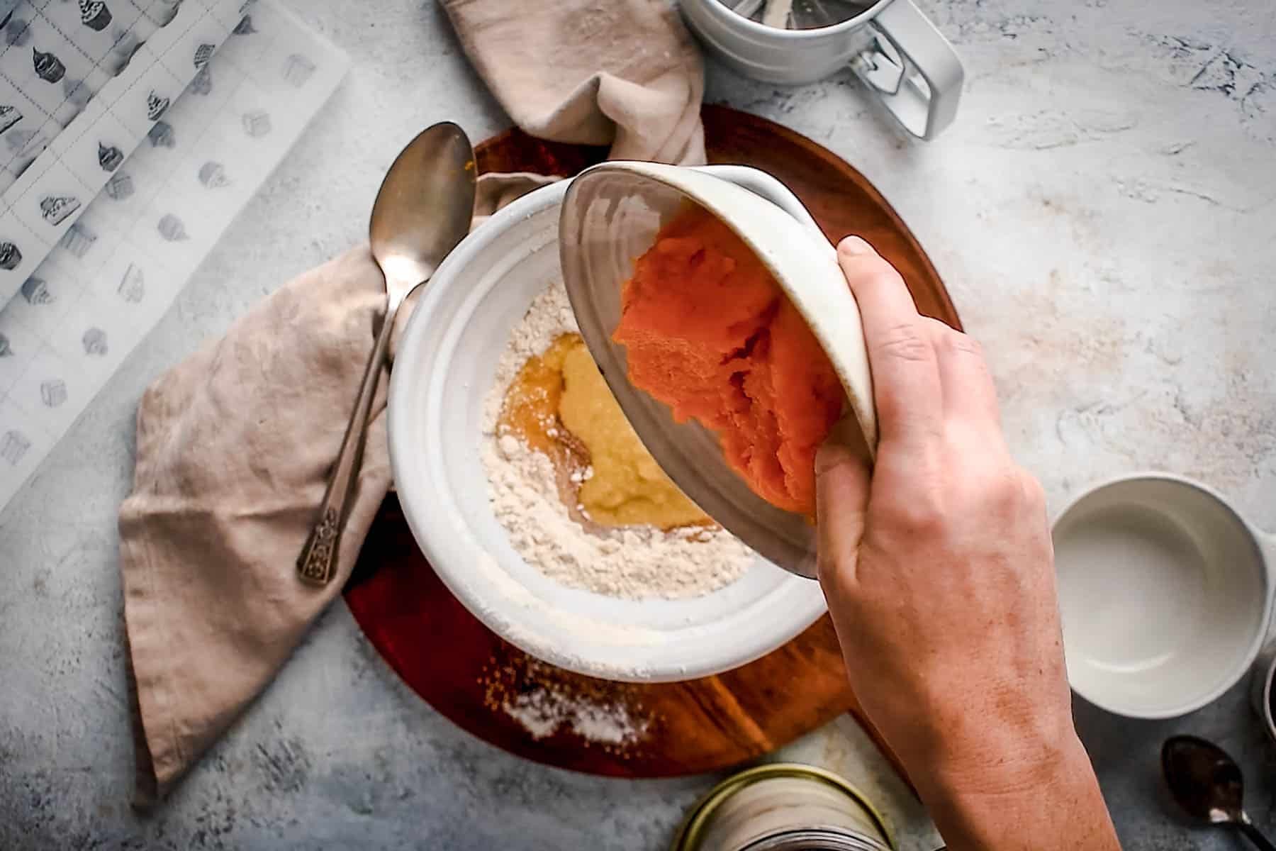 A person pouring a pumpkin puree into a bowl containing flour and other ingredients.