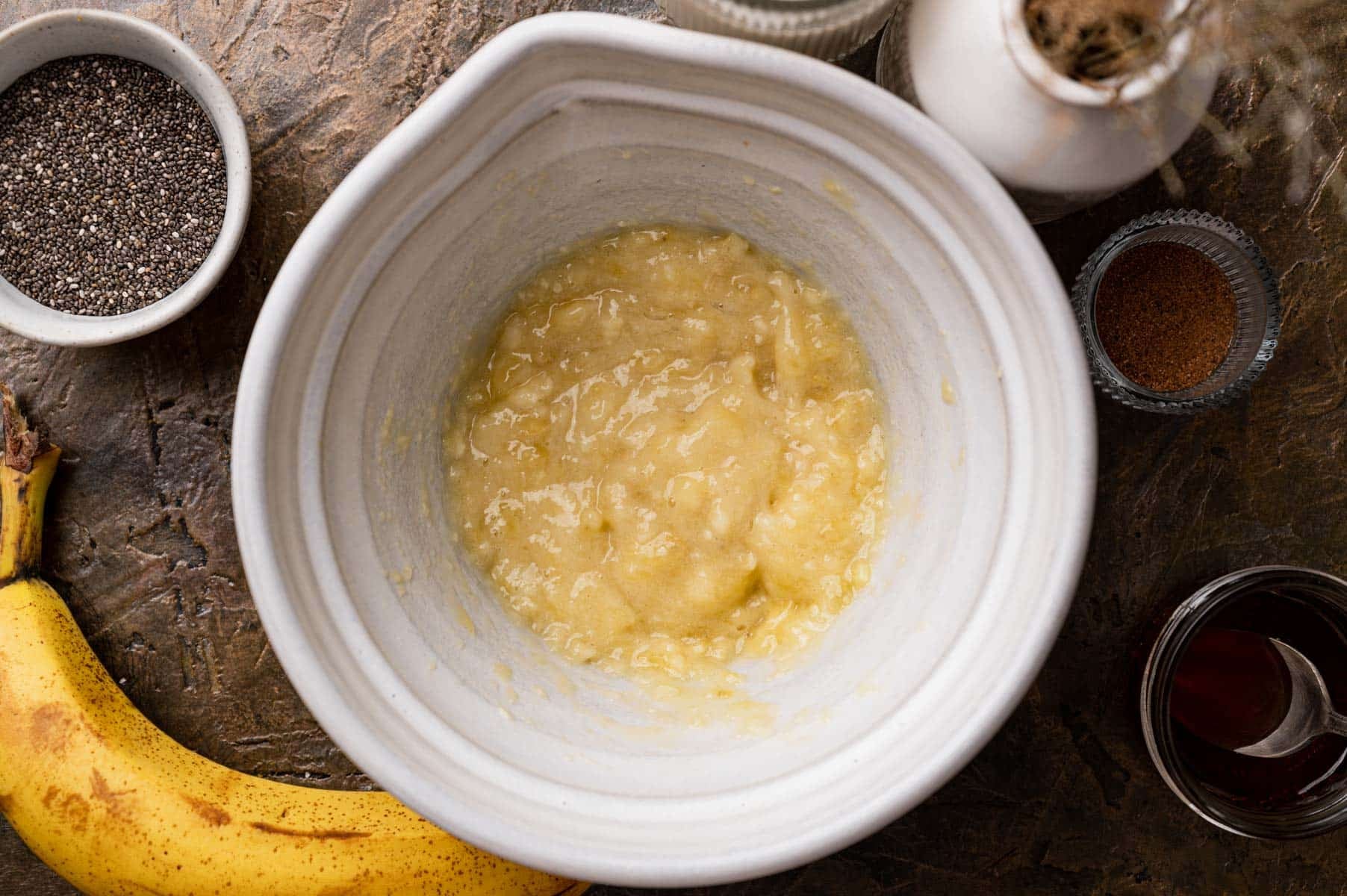 Mashed bananas in a bowl surrounded by a whole banana, chia seeds, cinnamon, a white jar, and a small glass of syrup on a textured surface.