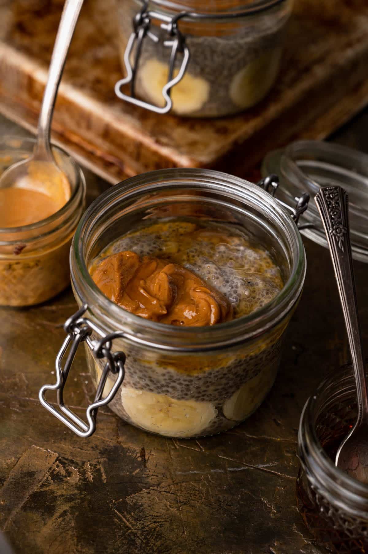 A jar of chia pudding topped with peanut butter and banana slices, with a spoon and a small bowl of peanut butter beside it.