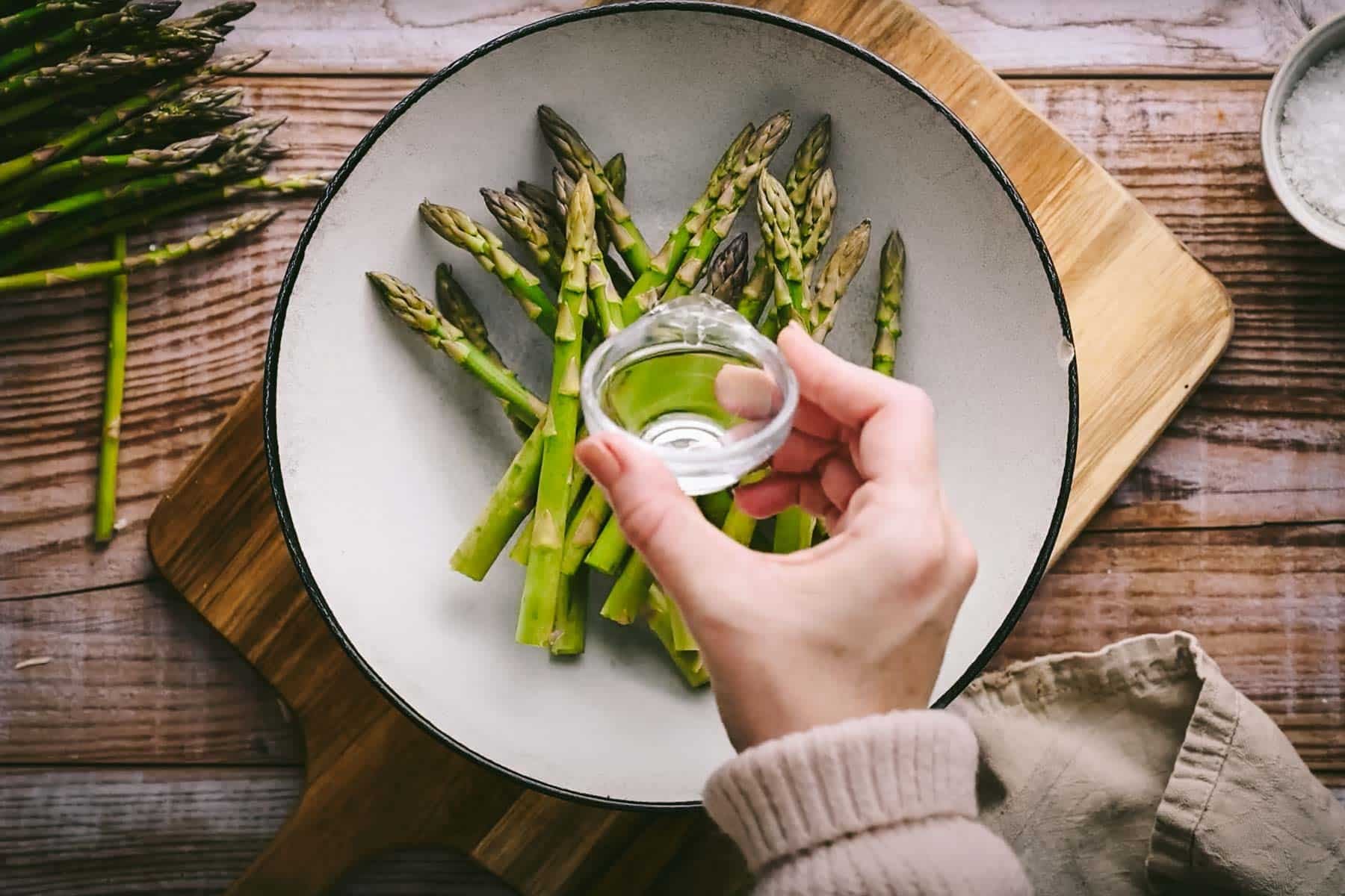 A hand pours water onto a bowl of fresh asparagus on a wooden cutting board.