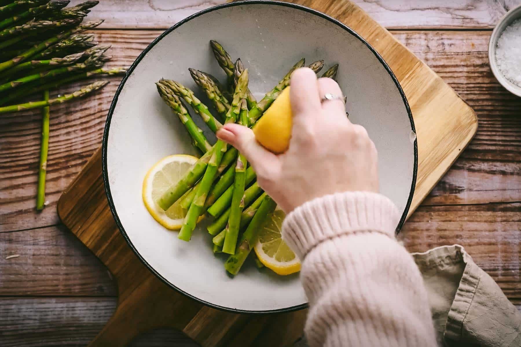 A hand squeezing a lemon over a dish of fresh asparagus spears arranged on a white plate with lemon slices, set on a wooden cutting board.