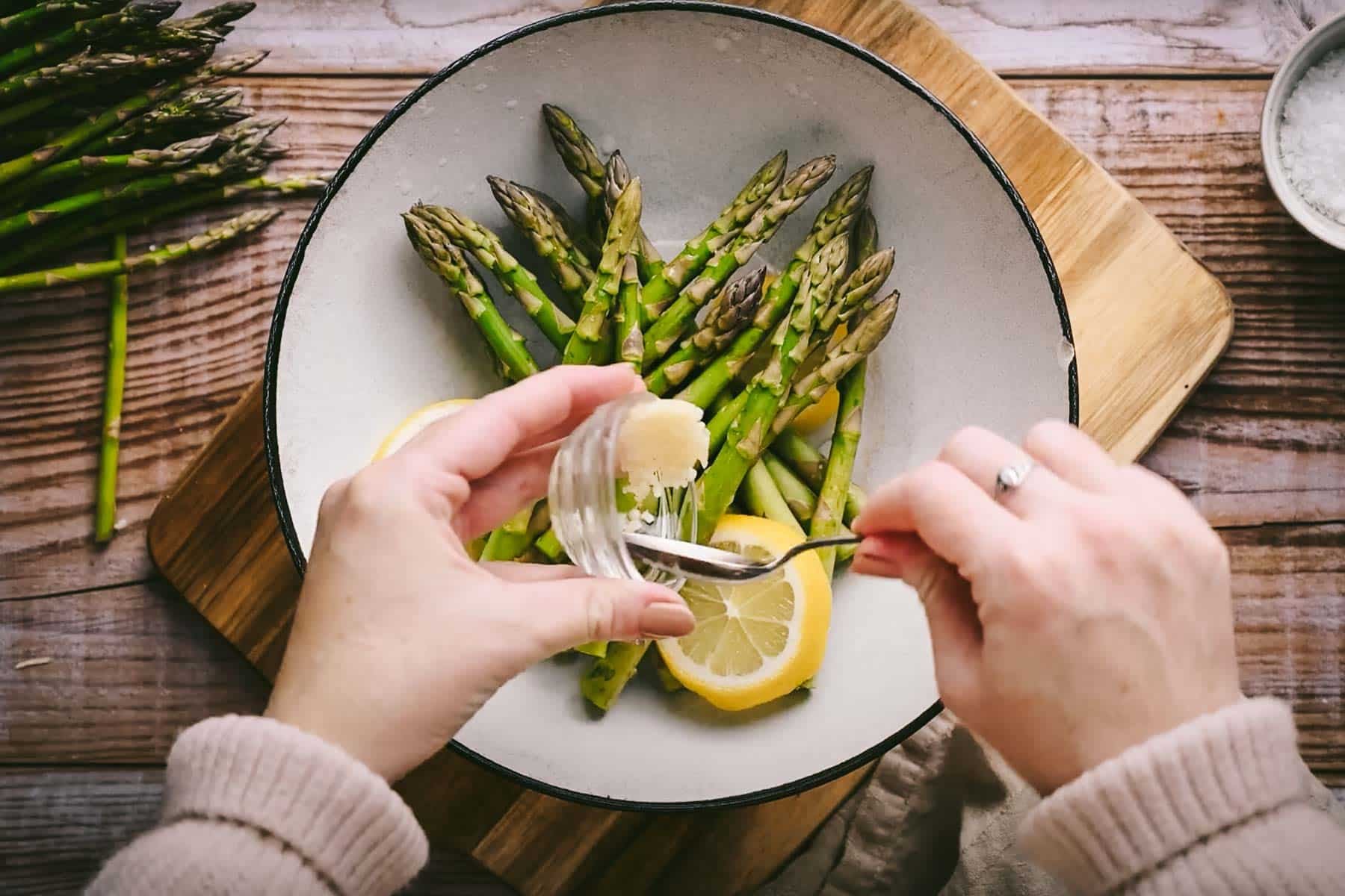 Person adding minced garlic over asparagus and lemon slices in a white bowl on a wooden countertop.