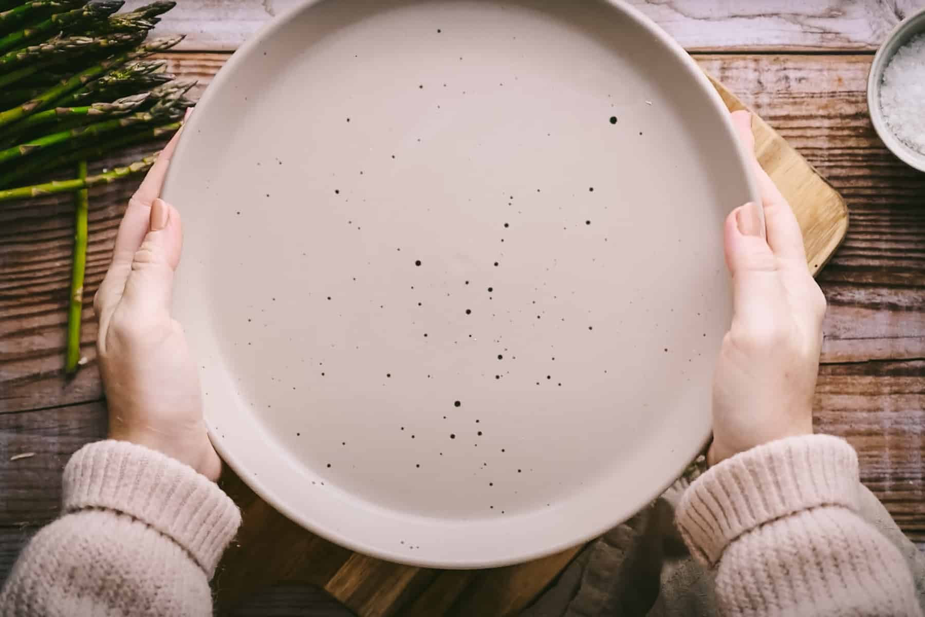 Hands holding an empty, round beige plate with black speckles, placed over plate of asparagus.