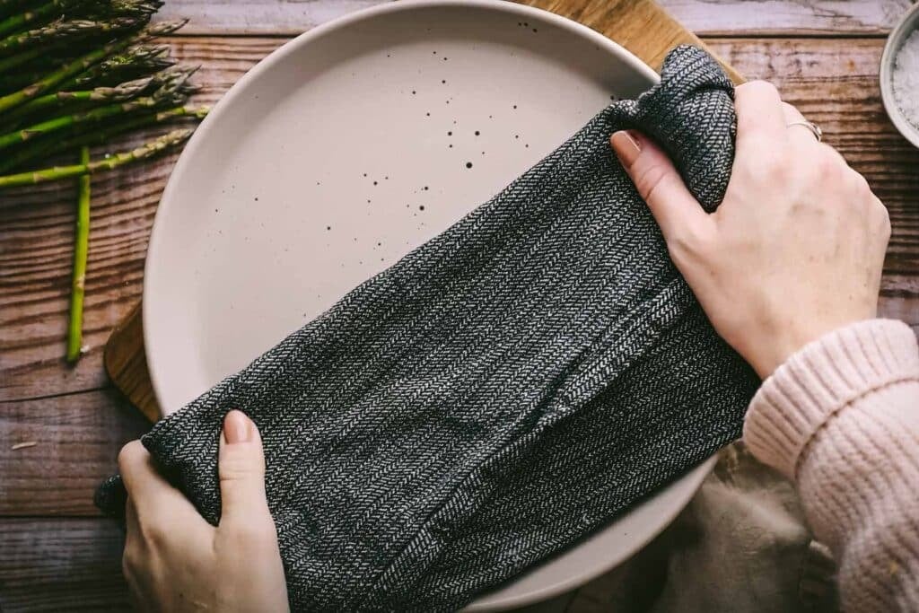 Hands placing a dark cloth napkin on a plate covering asparagus.