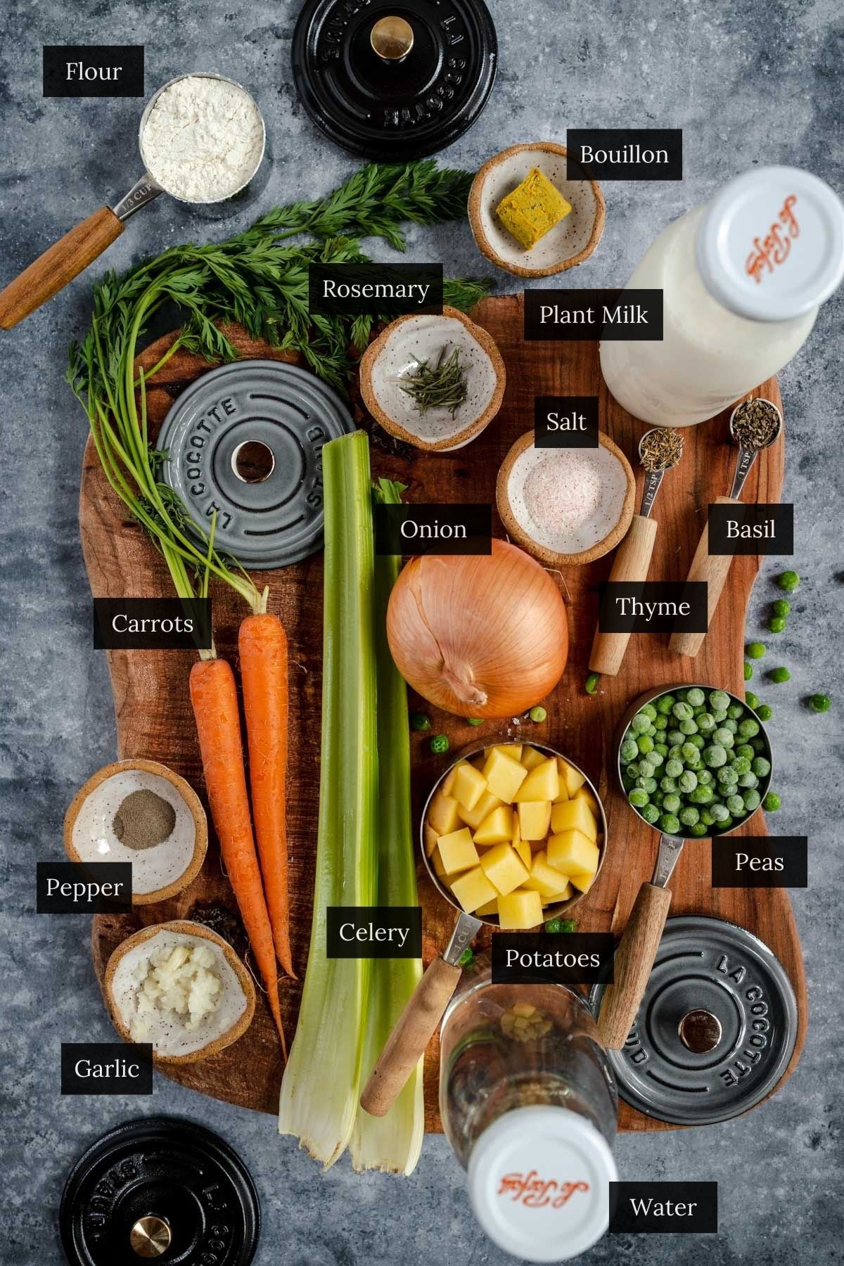 A wooden board artfully displays ingredients for mini pot pies: flour, rosemary, bouillon, plant milk, salt, basil, carrots, onion, thyme, celery, potatoes, peas, pepper, garlic, and water arranged neatly.