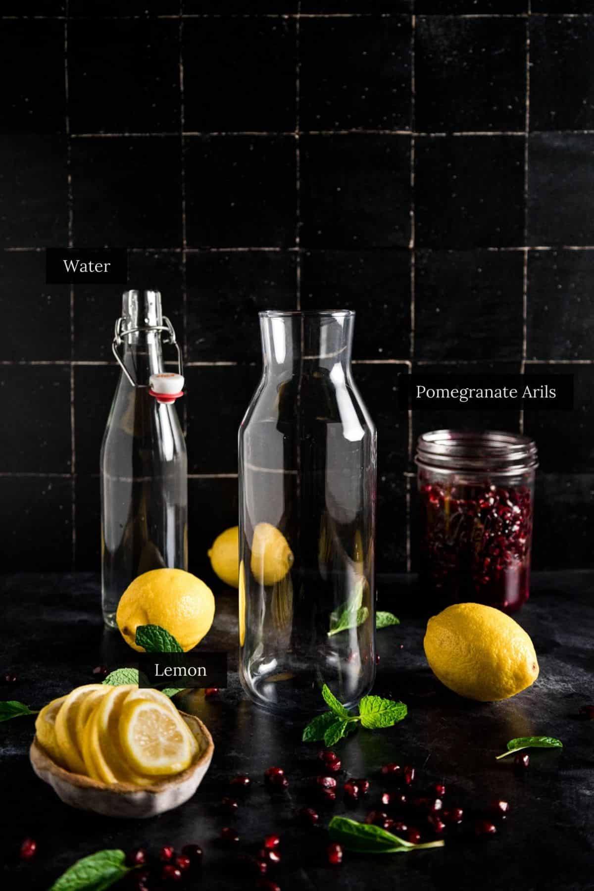 Glass bottles, a jar with pomegranate arils, sliced lemons, whole lemons, and mint leaves are arranged on a dark surface with a tiled background.