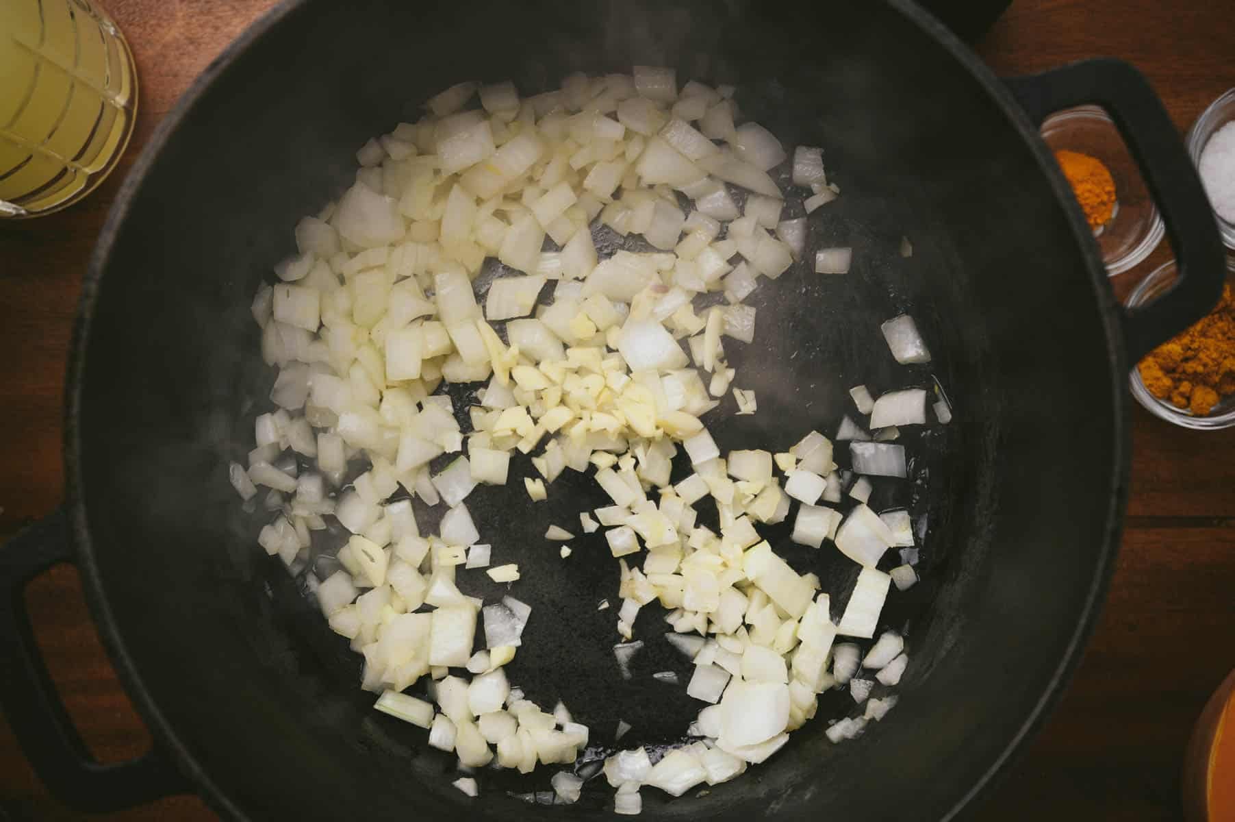 Diced onions sautéing in a black pot, surrounded by small bowls of spices and oil on a wooden surface.