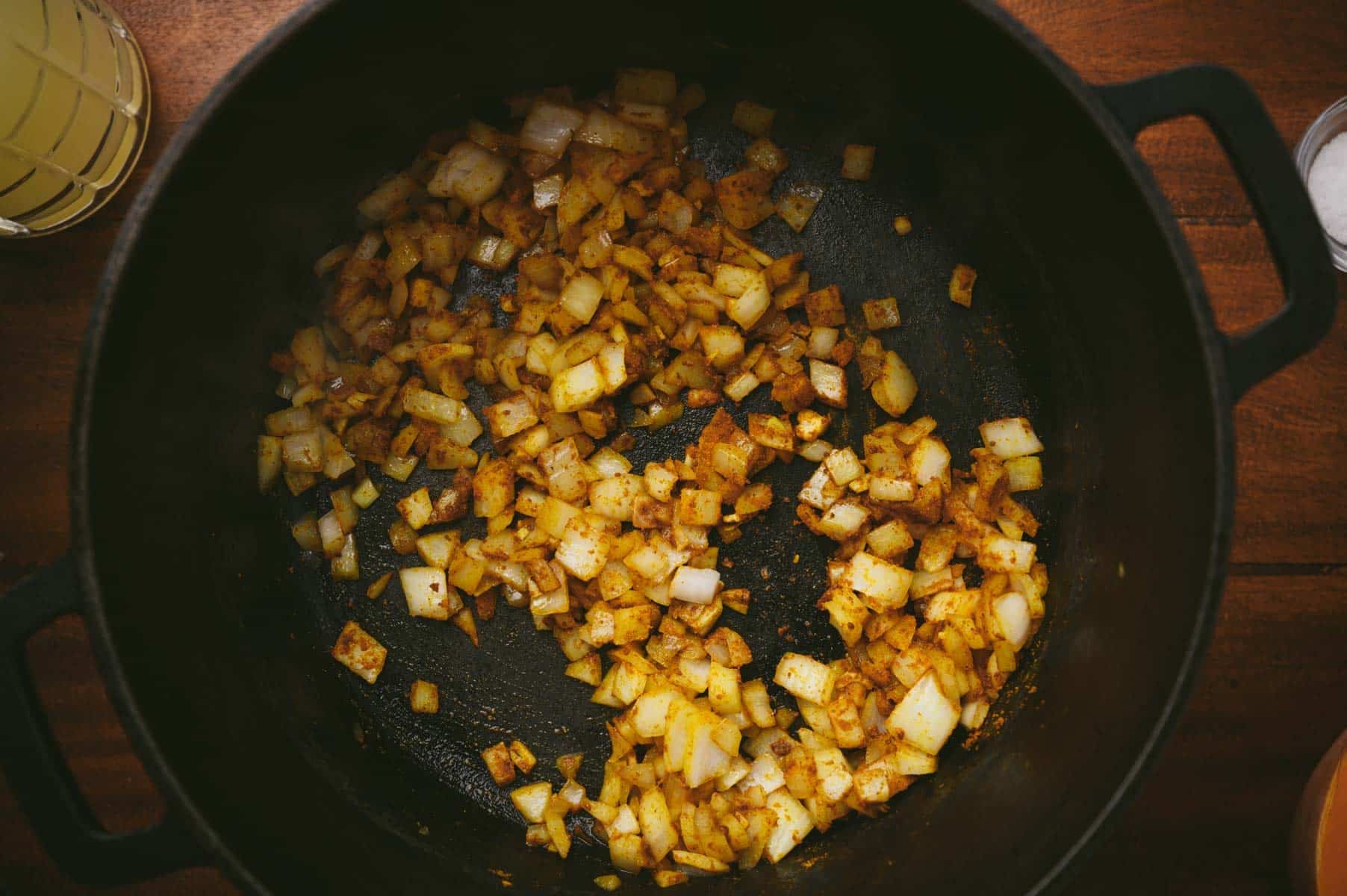 Chopped onions sautéing in a dark skillet with seasoning, on a wooden surface.