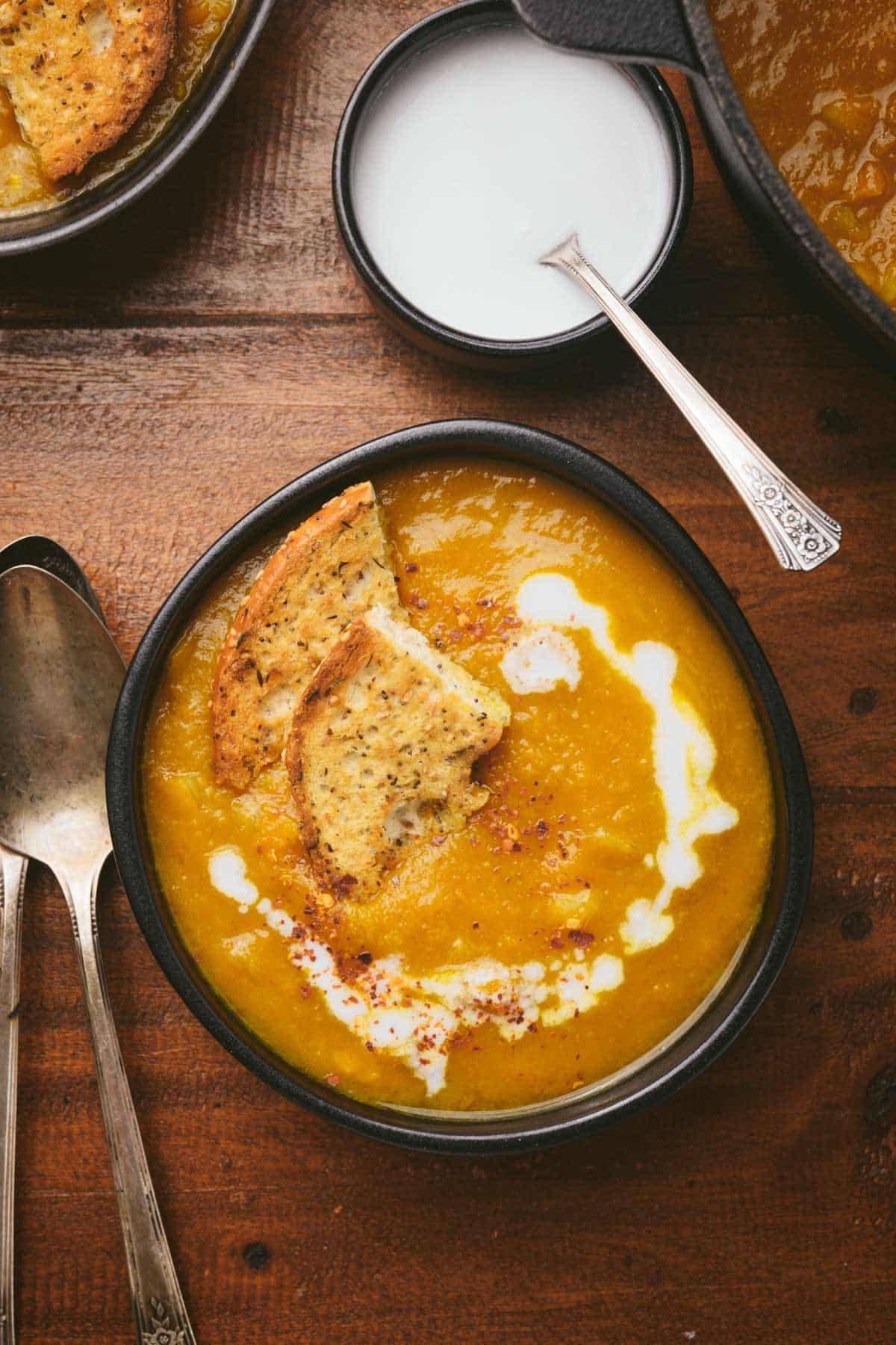 A bowl of pumpkin soup topped with coconut cream and garnished with sesame crackers on a wooden table.