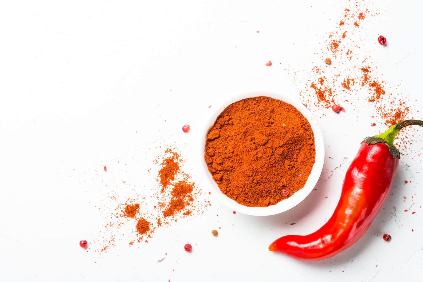 A bowl of ground chili powder and a whole red chili pepper on a white surface, with scattered pepper flakes.