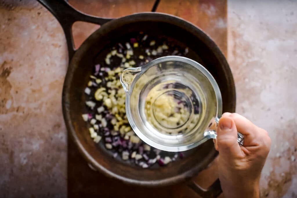 A hand pours liquid from a glass measuring cup into a pot containing chopped onions.