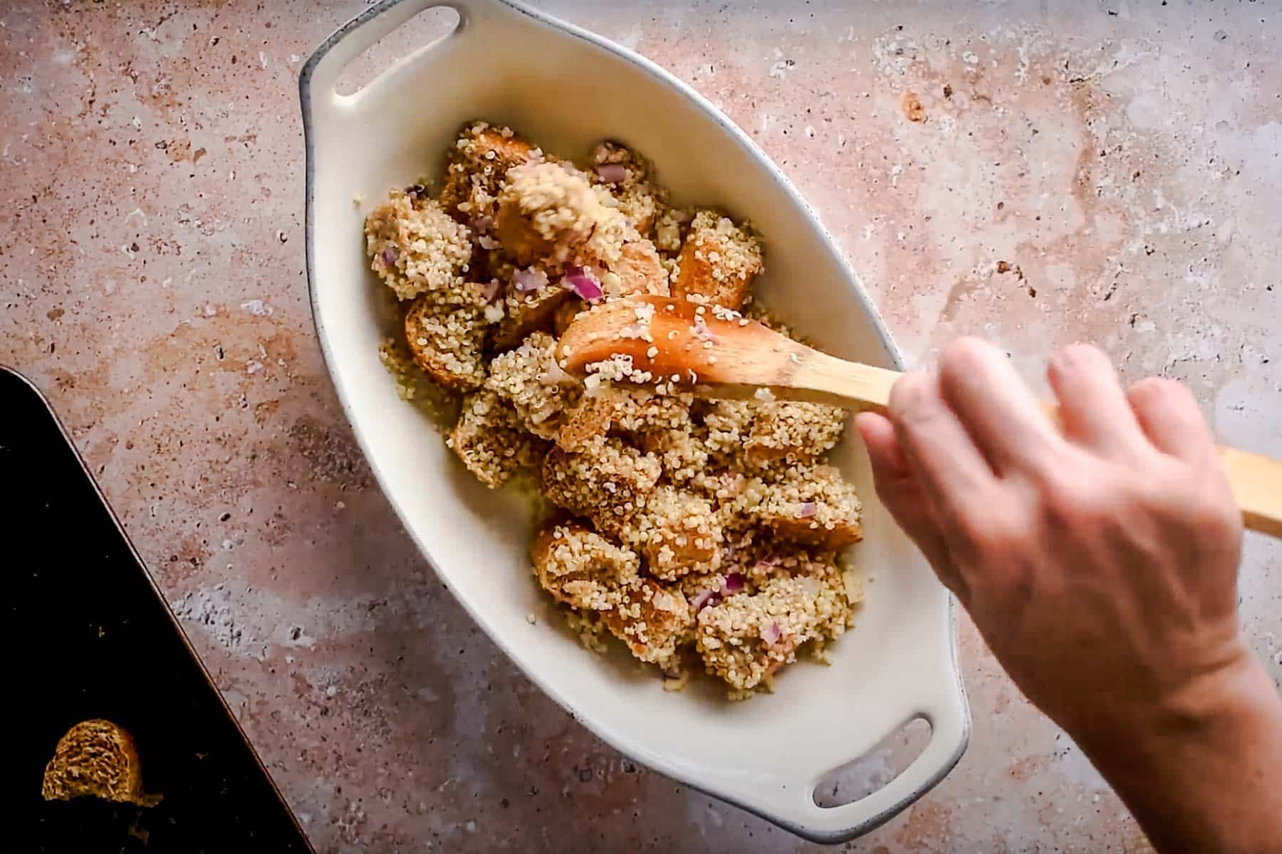 A person mixes seasoned vegan stuffing with quinoa and chopped onions in a white baking dish using a wooden spoon.