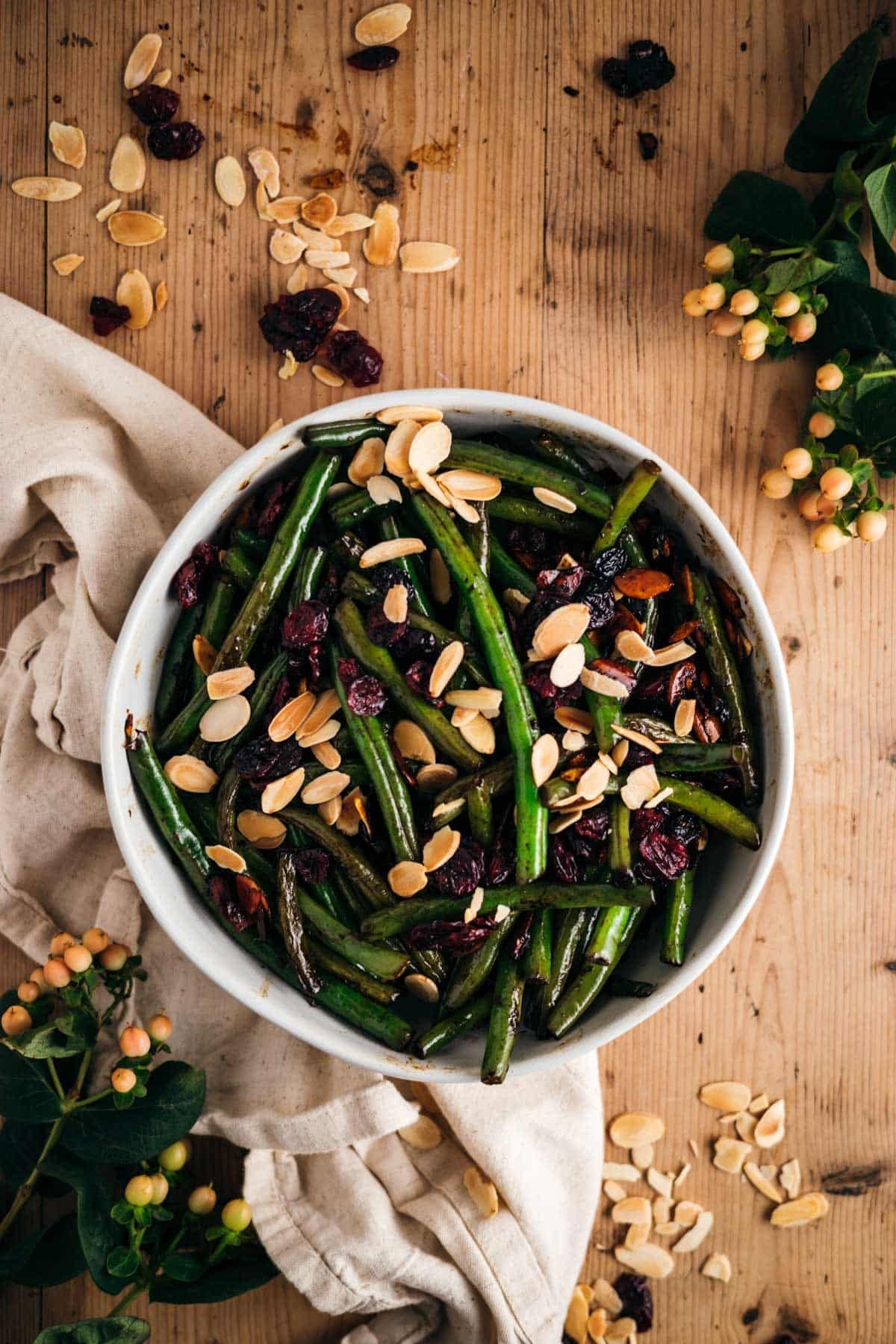 A bowl of green beans topped with sliced almonds and dried cranberries on a wooden table, surrounded by scattered nuts, berries, and foliage.