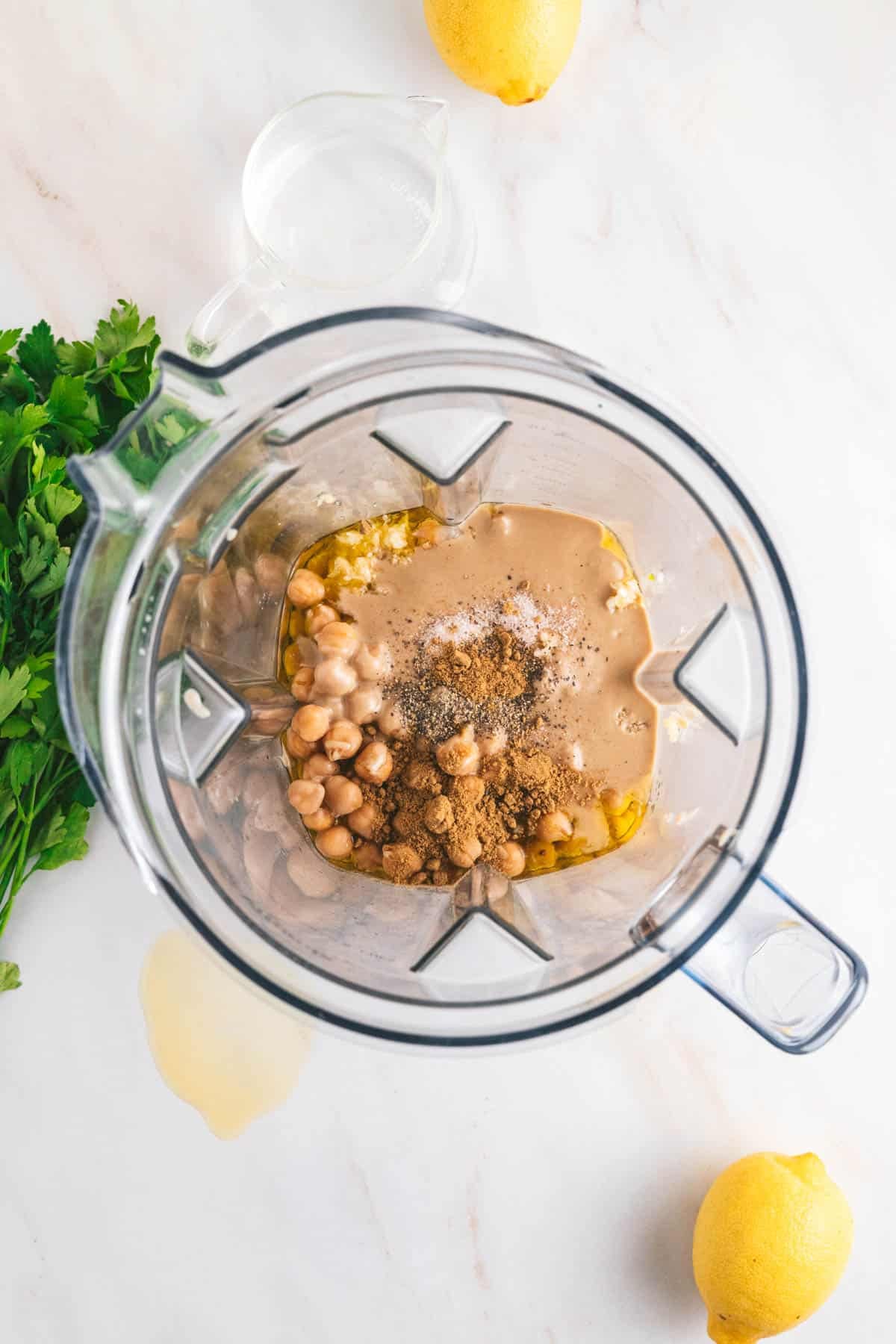 Ingredients for hummus in a blender, including chickpeas, tahini, olive oil, garlic, and spices. Lemons and parsley are nearby on the countertop.
