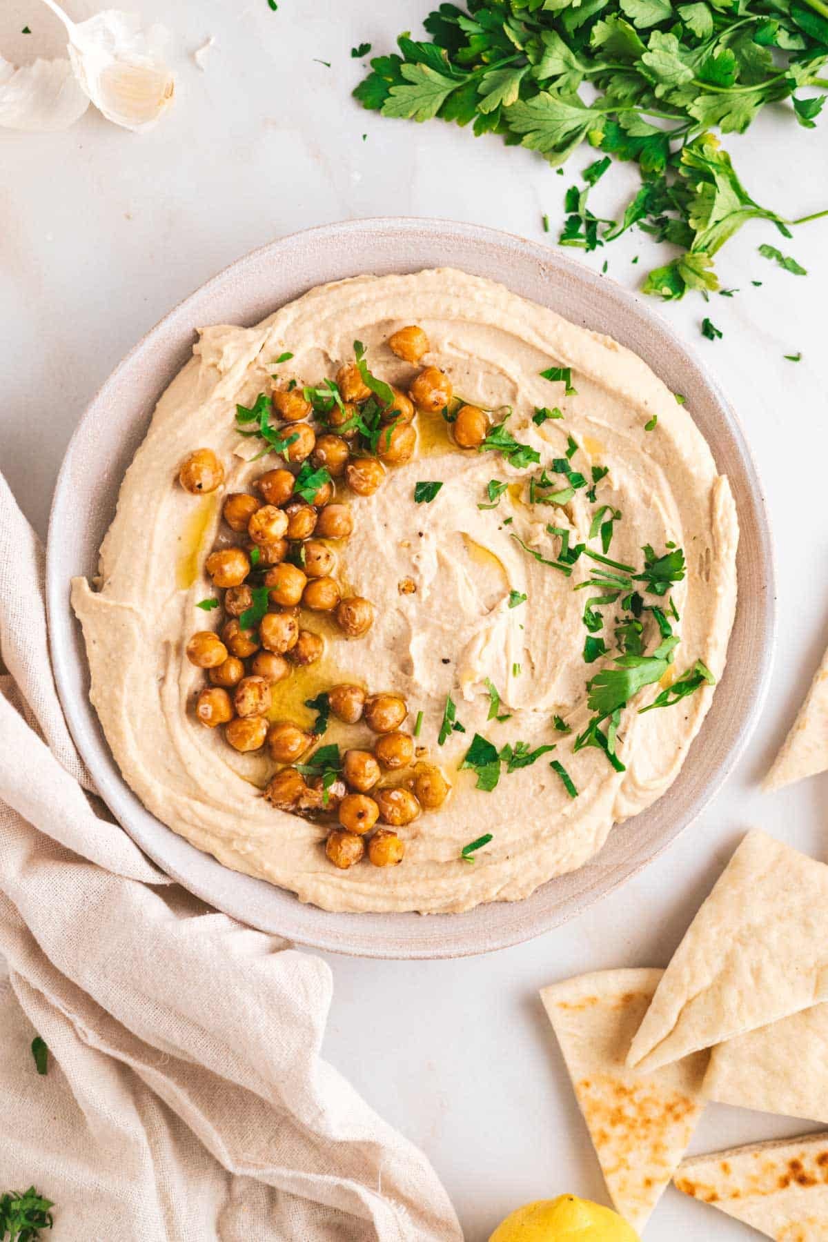 A plate of hummus topped with roasted chickpeas and parsley, surrounded by pita bread slices, garlic, and fresh herbs on a white surface.