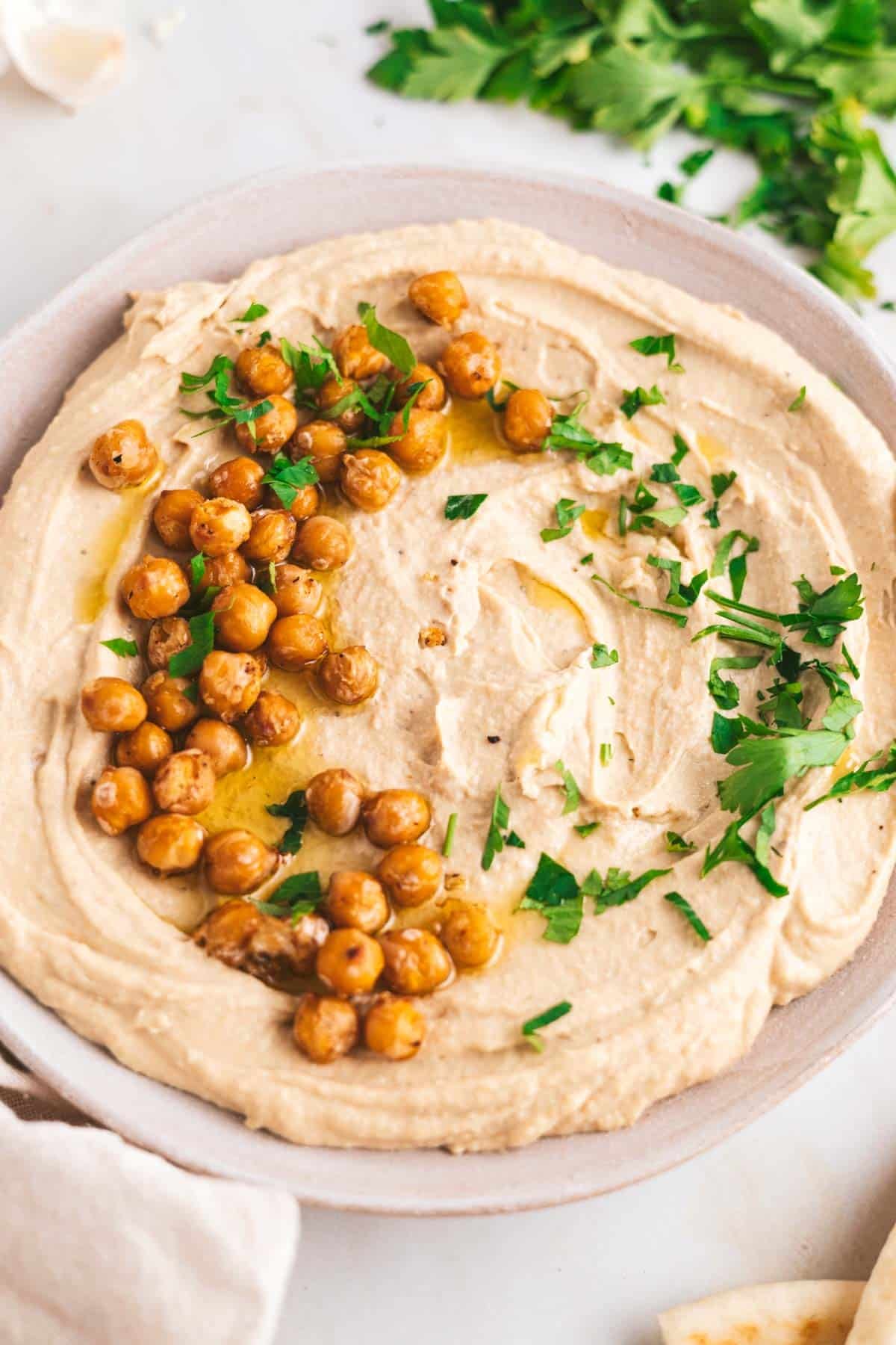 A bowl of hummus topped with roasted chickpeas, olive oil, and chopped parsley on a white table.