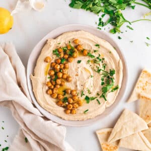 A bowl of hummus garnished with roasted chickpeas and parsley, surrounded by pita bread, fresh parsley, a lemon, and a beige cloth on a white surface.