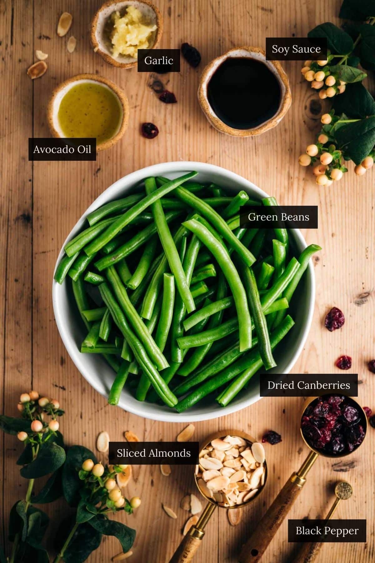 A bowl of green beans, accompanied by avocado oil, soy sauce, garlic, sliced almonds, black pepper, and dried cranberries sits elegantly on a wooden table. The addition of oat milk adds a surprising twist to this colorful ensemble.