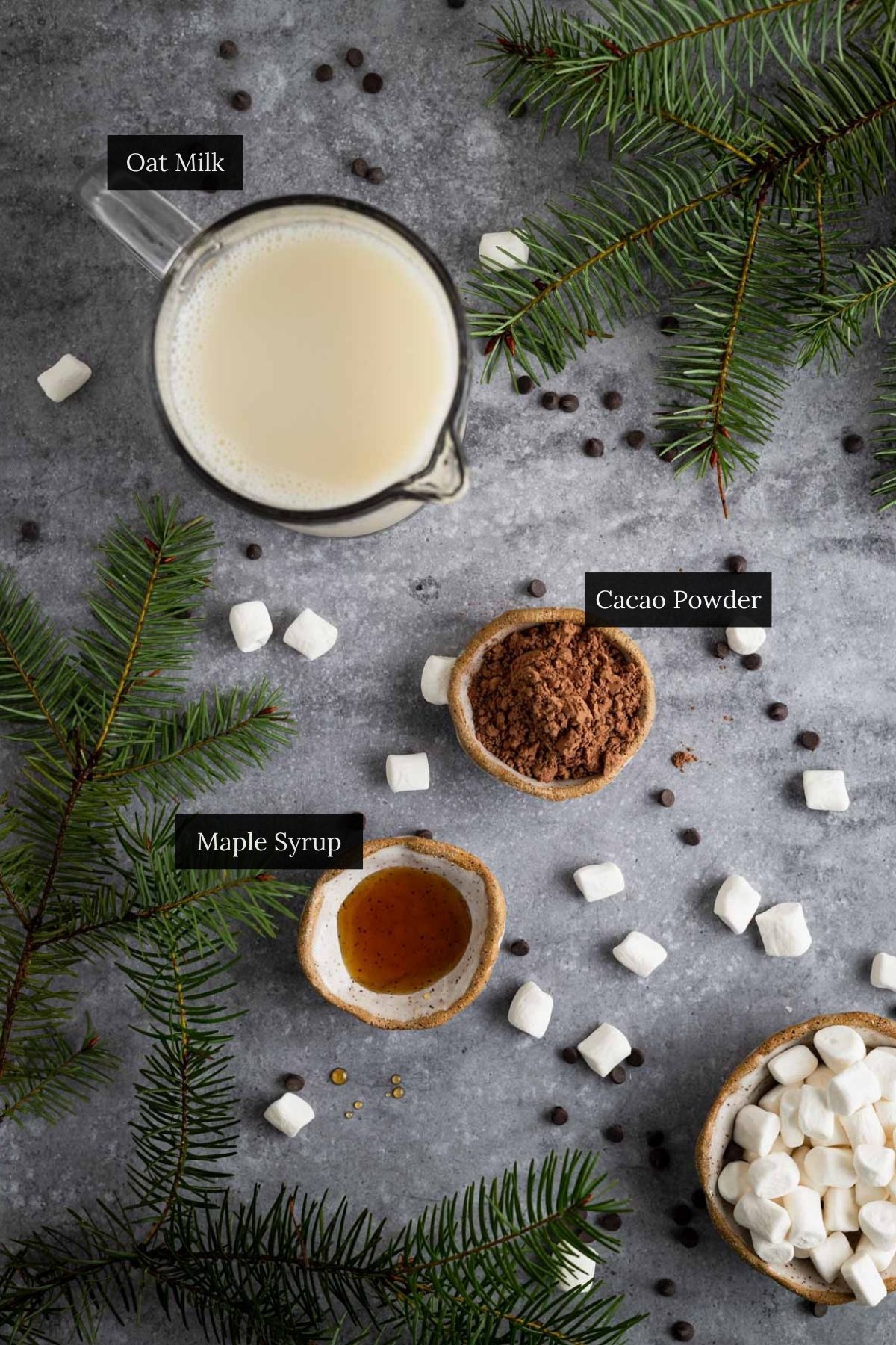 A cozy scene features Oat Milk Hot Chocolate essentials: oat milk in a glass jug, cacao powder and maple syrup in bowls, marshmallows, and green fir branches adorning the gray surface.
