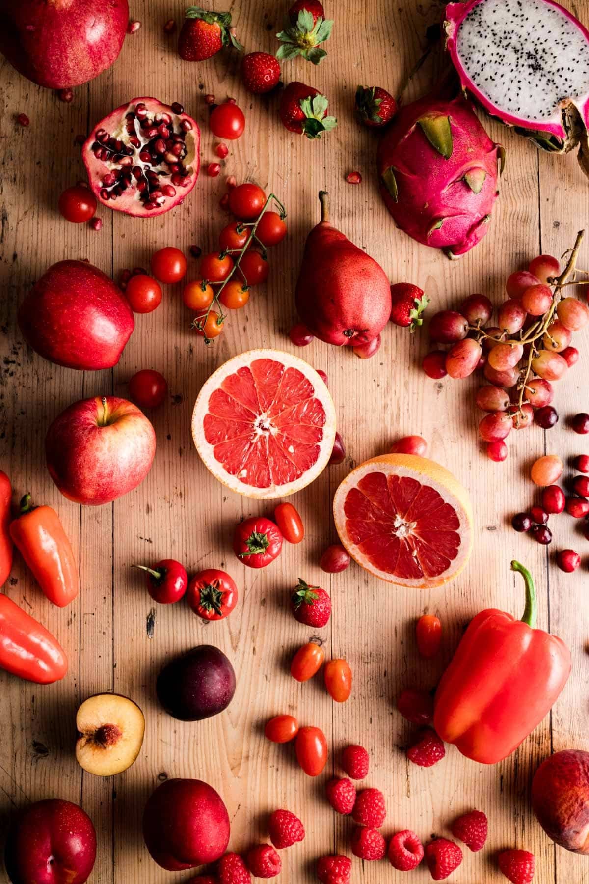 A variety of red fruits and vegetables, including grapefruits, strawberries, tomatoes, pomegranates, and bell peppers, are scattered on a wooden surface.