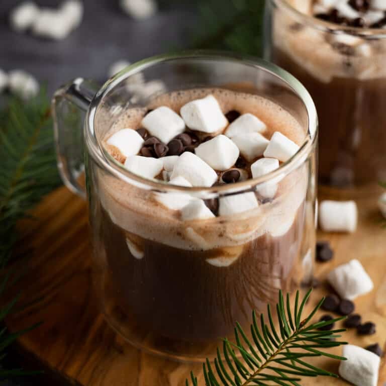 A clear mug of hot chocolate topped with mini marshmallows and chocolate chips, surrounded by pine branches and more marshmallows on a wooden surface.
