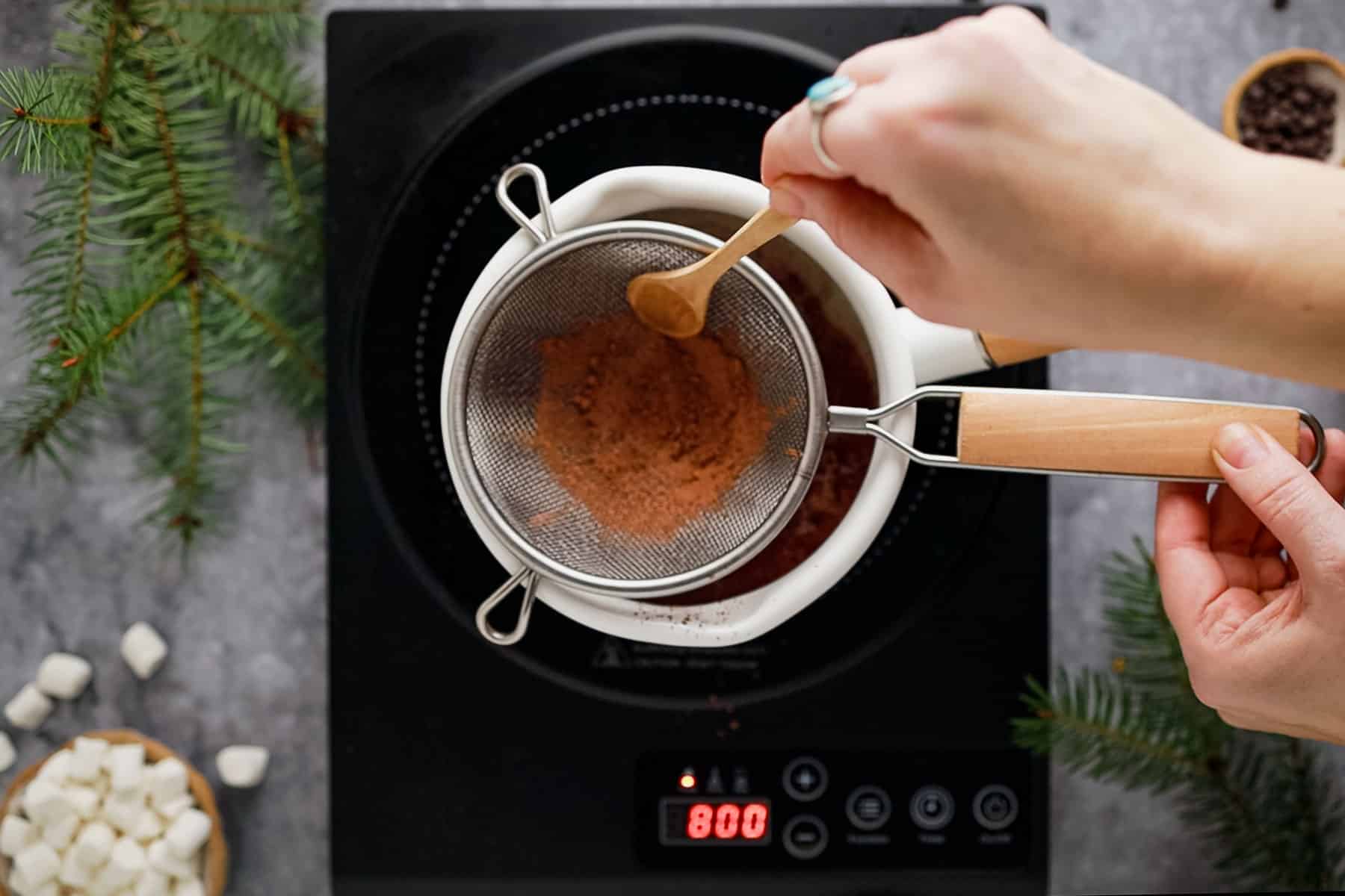 Person sifts cocoa powder into a bowl on an induction stove, surrounded by pine branches and marshmallows.