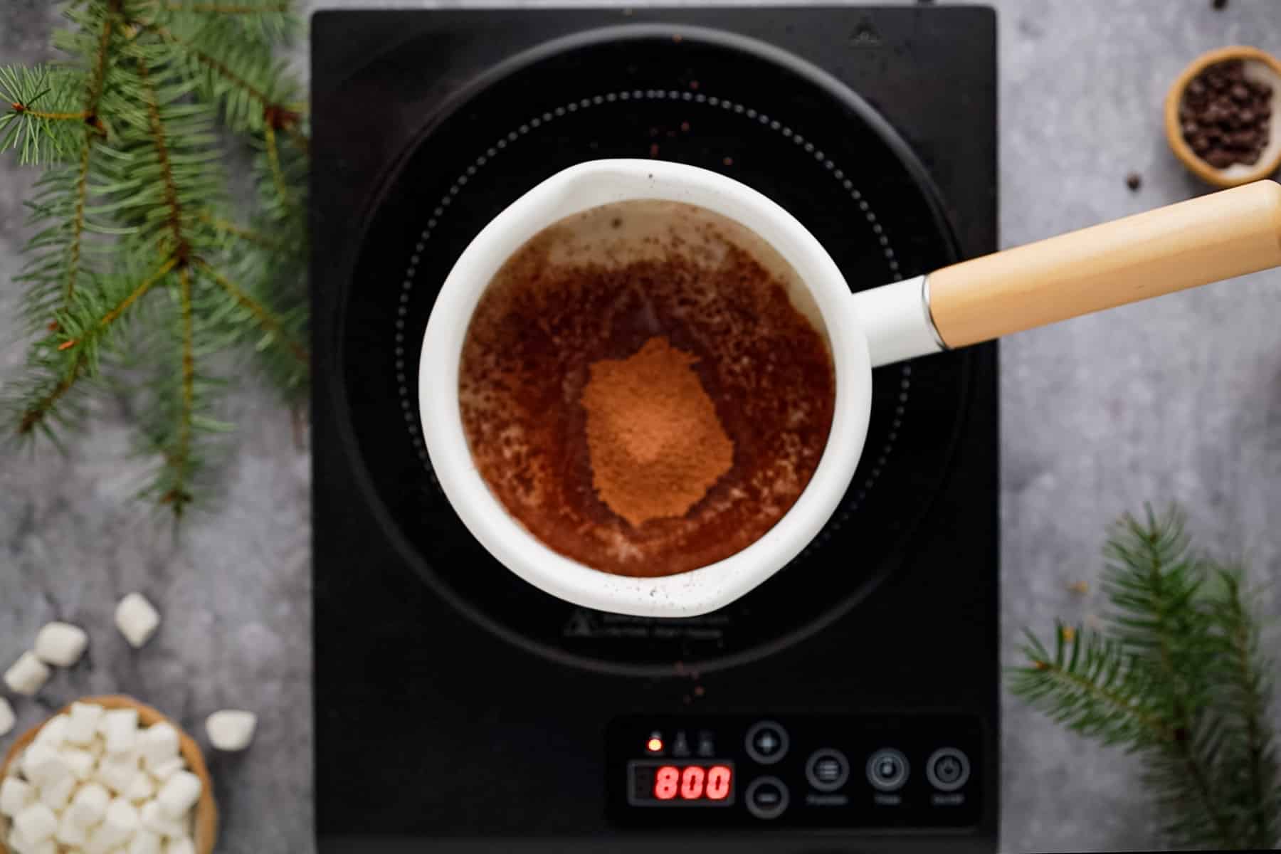 A pot with cocoa on an induction stove, surrounded by marshmallows and greenery.