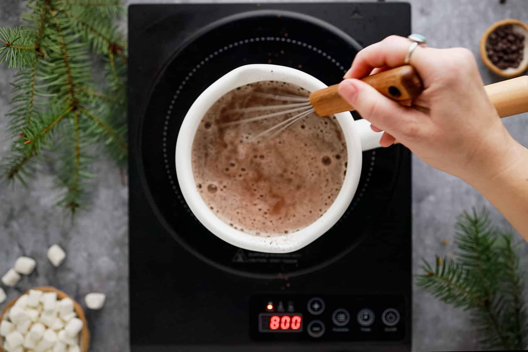 Overhead view of a person whisking hot chocolate in a saucepan on an induction cooktop. Marshmallows and pine branches are nearby.