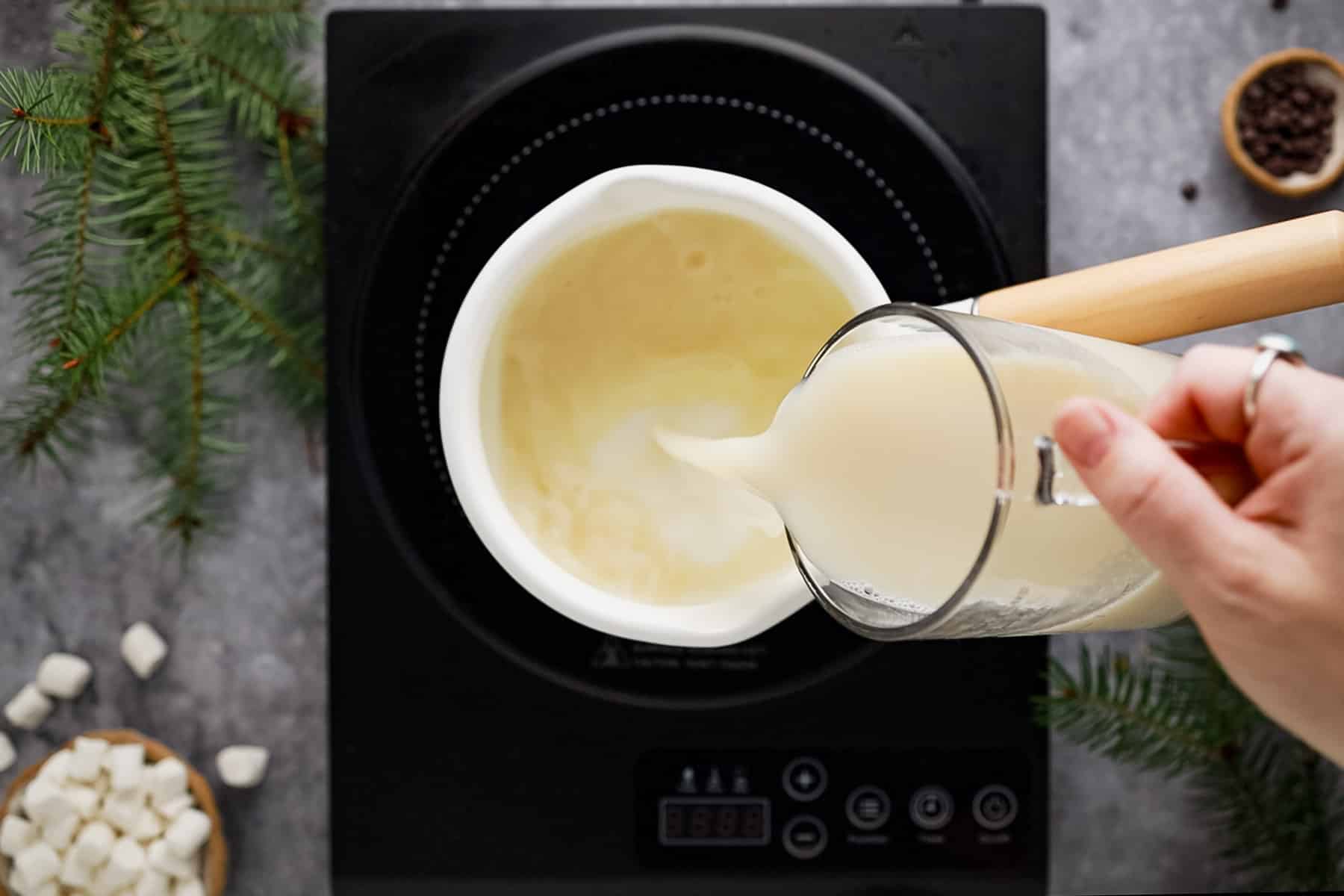 Person pouring milk into a white saucepan on an induction stove, surrounded by evergreen branches and marshmallows.