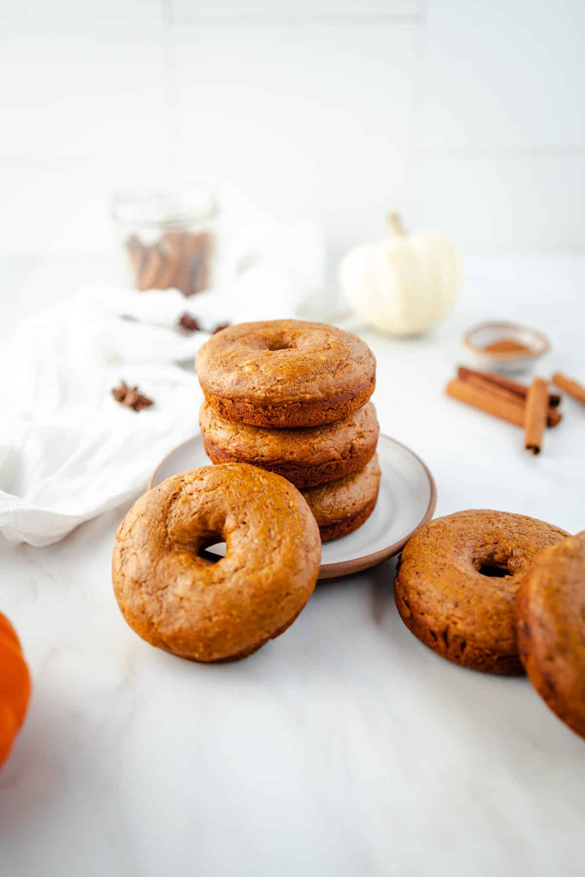 A stack of pumpkin spice donuts on a plate is surrounded by whole donuts, cinnamon sticks, and a white pumpkin on a marble surface.