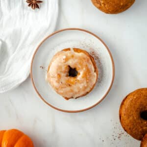 A glazed donut sits on a white plate surrounded by pumpkin spice donuts, with a scattering of cinnamon adorning the marble surface.