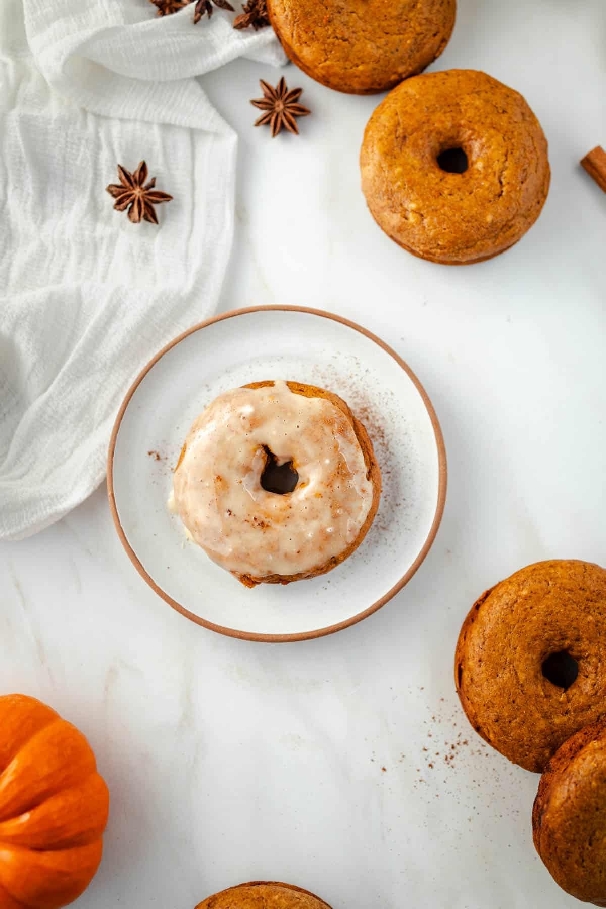 A glazed donut on a white plate is surrounded by pumpkin spice donuts, star anise, and a small pumpkin on a marble surface.