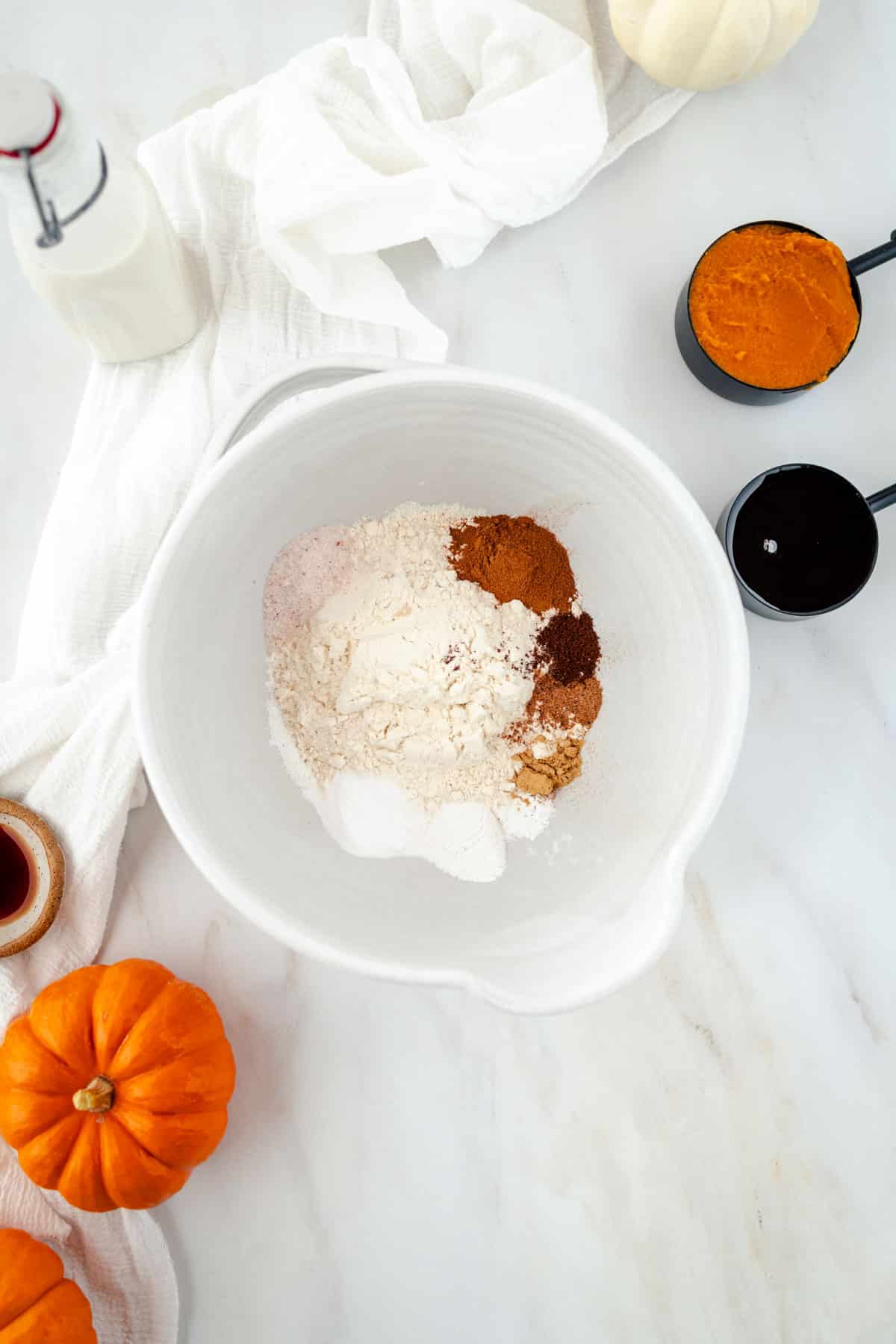 White mixing bowl with flour and spices on a white surface, surrounded by small pumpkins, a bottle of milk, and a cup of pumpkin puree.