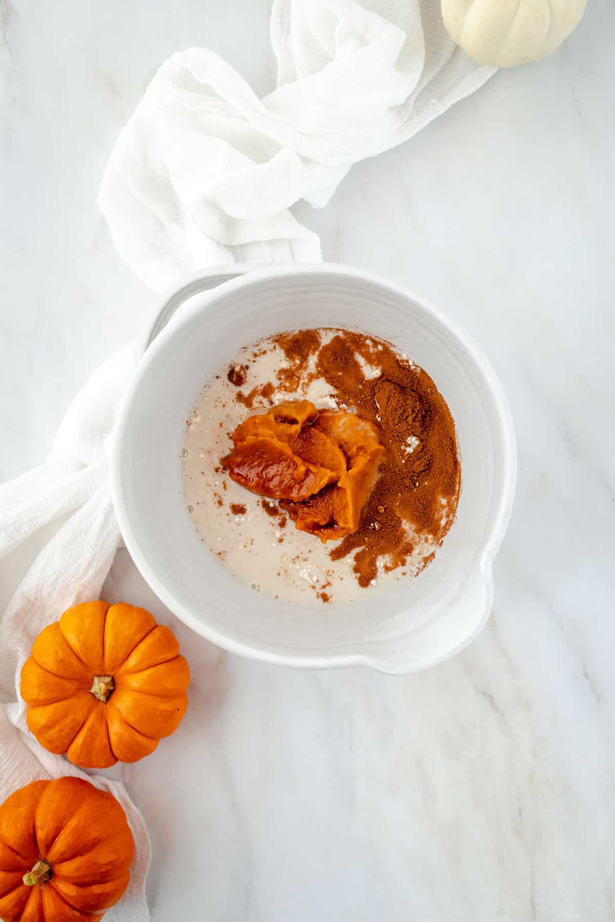 White bowl with pumpkin puree, spices, and other ingredients on a marble surface. Two small pumpkins and a white cloth are beside the bowl.