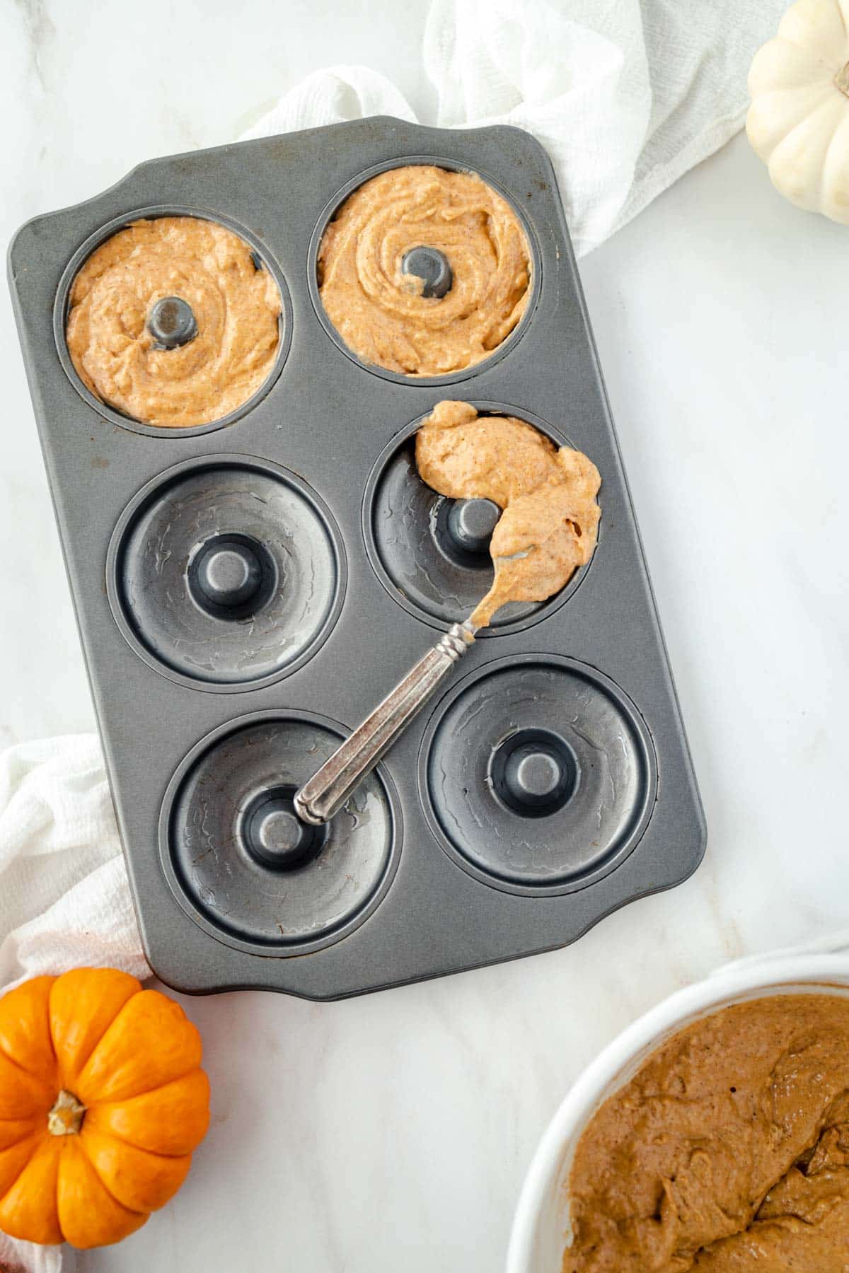 Batter-filled donut pan with a spoon, surrounded by small pumpkins on a white countertop.
