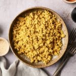 A bowl of scrambled tofu on a textured surface, surrounded by small dishes and forks.