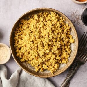 A bowl of scrambled tofu on a textured surface, surrounded by small dishes and forks.